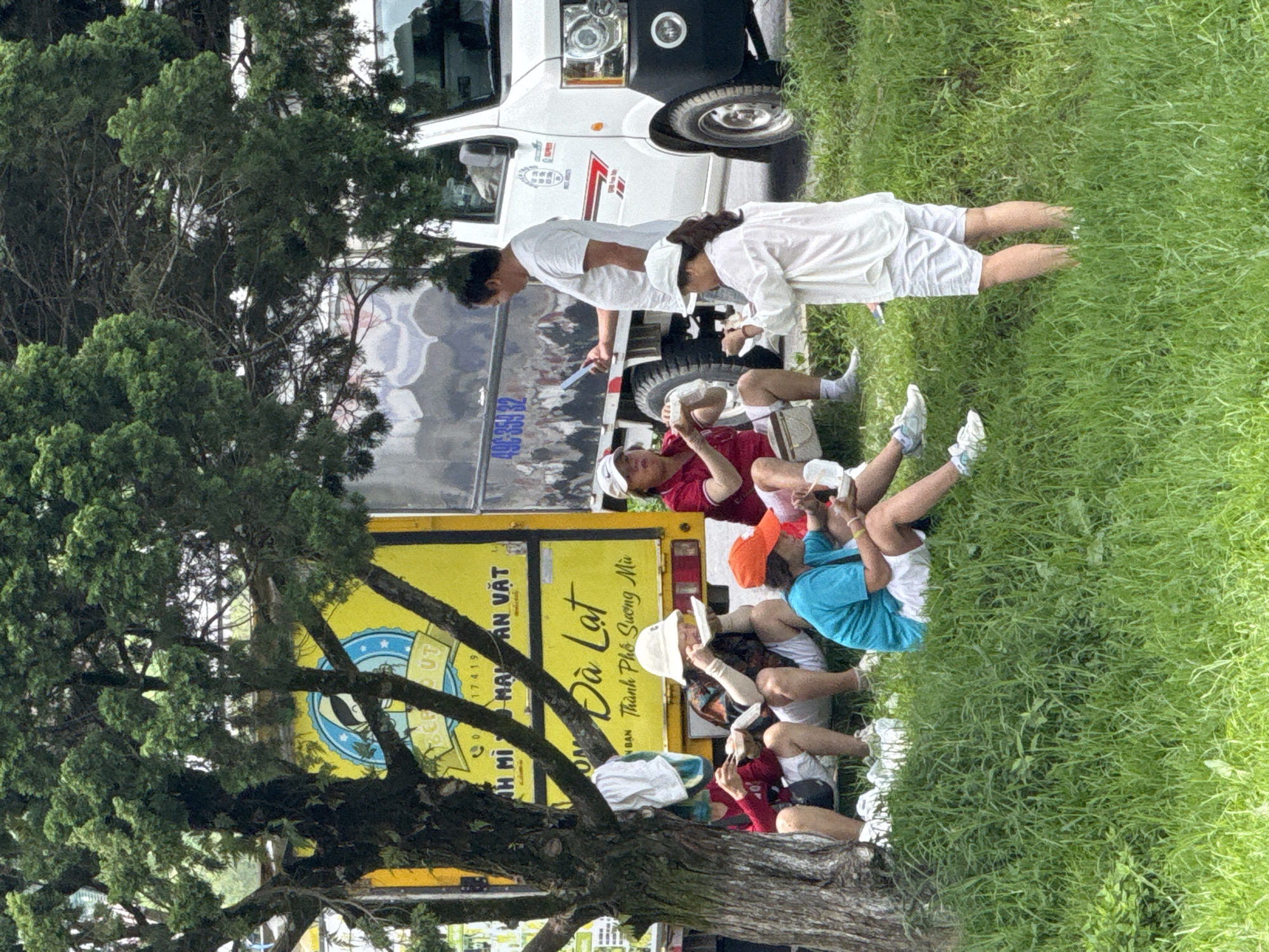 In Phường Xuân Hương - Đà Lạt, Vietnam, a group of individuals is gathered outdoors, seated and standing near a parked vehicle. A yellow truck with Vietnamese writing is visible on its side, featuring the words "Đà Lạt" and "Thành Phố Sương Mù." A white truck is parked next to it. Several people are eating from takeout containers. One woman in a white hat and patterned shirt is seated and eating. To her left, two individuals are also seated and eating. Another person in a red shirt and white shorts is seated and holding a food container. A person in a blue shirt and white shorts is seated on the grass. A man in a white t-shirt is standing and holding a mobile phone. A woman in a white top and white shorts is standing and appears to be interacting with her phone. The scene is set amidst lush green grass and under the canopy of a large tree.