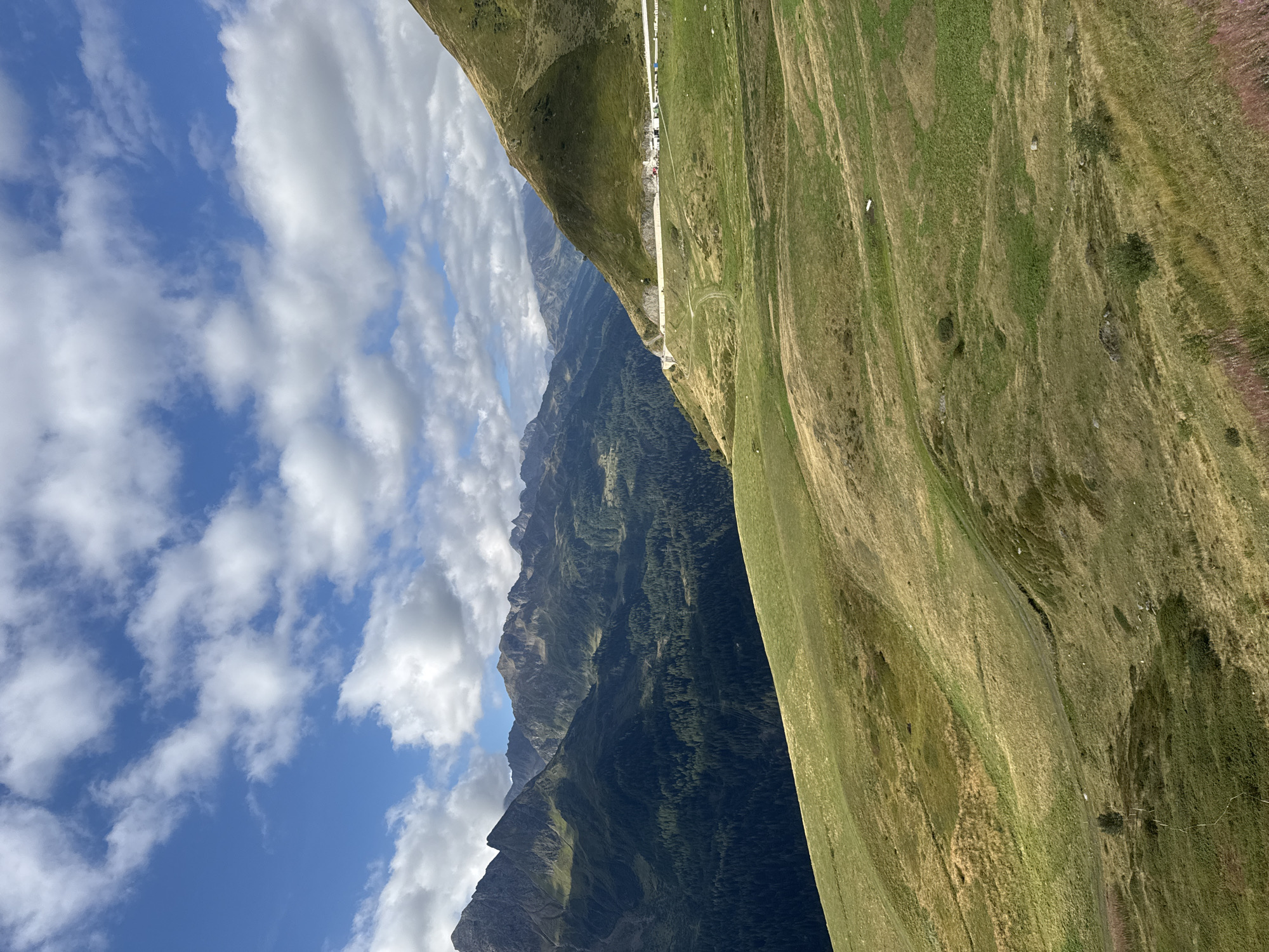 The image depicts a mountainous landscape in Airolo, Switzerland. The foreground features rolling green hills with patches of dry grass and scattered vegetation. A winding path is visible on the lower slopes. In the middle ground, a steep, grassy mountainside rises, with a road carved into its slope. White guardrails line the road, and several white buildings and vehicles are visible on or near it. The background is dominated by a range of rugged, dark mountains covered in dense green forest in the lower sections and rocky, barren peaks higher up. The sky is a clear blue with scattered white cumulus clouds.