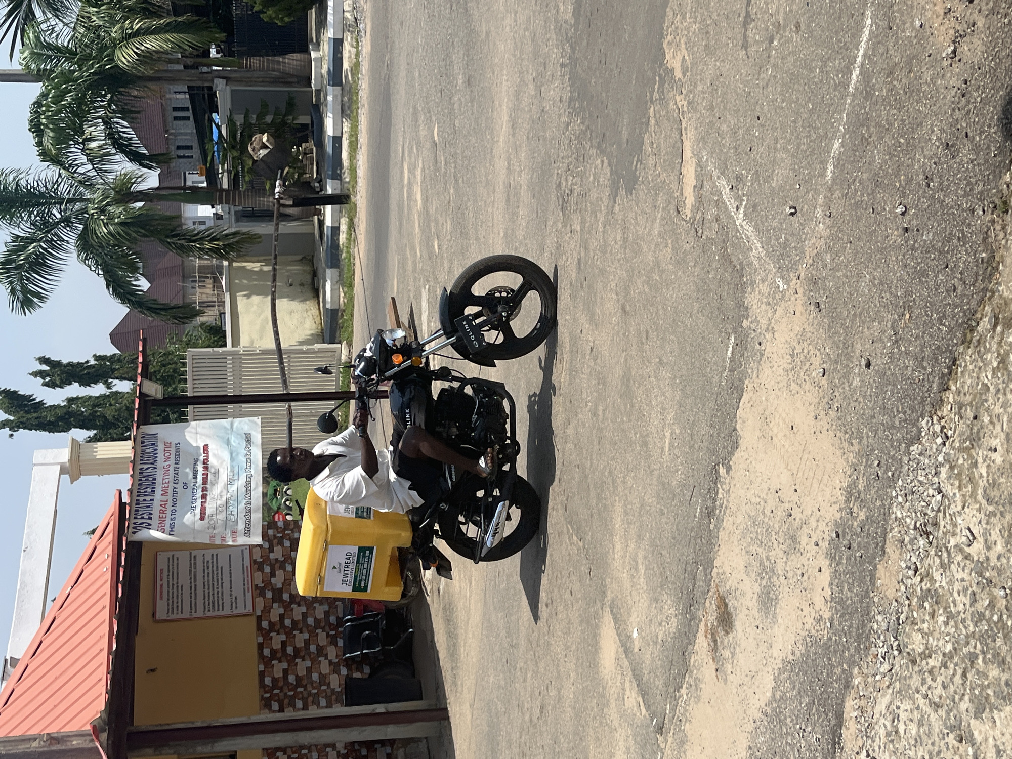 In Abuja, Nigeria, a man rides a motorcycle with a yellow cargo box on the back. He is wearing a white t-shirt and dark shorts. The motorcycle has a "QOLINK" logo on the front fender. Behind him, a sign for the "GOS ESTATE RESIDENTS ASSOCIATION" is posted on a building. Palm trees and residential buildings are visible in the background. The road is paved with asphalt and shows signs of wear.