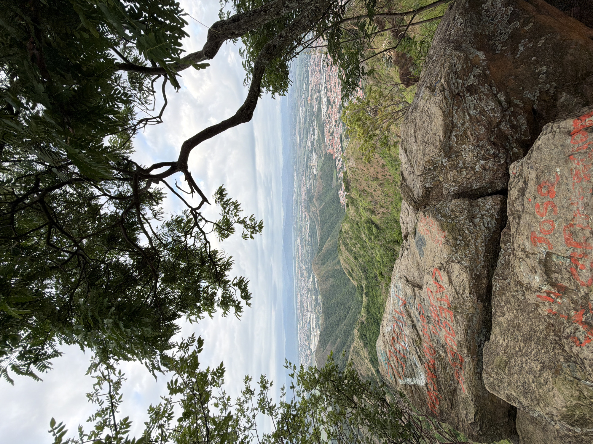 The image depicts a panoramic view from a rocky outcrop, likely on a mountainside overlooking a city. In the foreground, large, weathered rocks are visible, some with red graffiti. The terrain slopes downwards, covered in lush green vegetation, transitioning into rolling hills. Further in the distance, a sprawling city is spread out, characterized by numerous buildings with red tile roofs, interspersed with industrial structures and green spaces. The city extends towards a hazy horizon where mountains meet the sky under a cloudy, overcast day. Trees with dark green foliage frame the scene from the top and sides, suggesting a natural environment above the urban area. The overall impression is of a high vantage point offering a scenic vista of a populated valley in El Cambur, Venezuela.