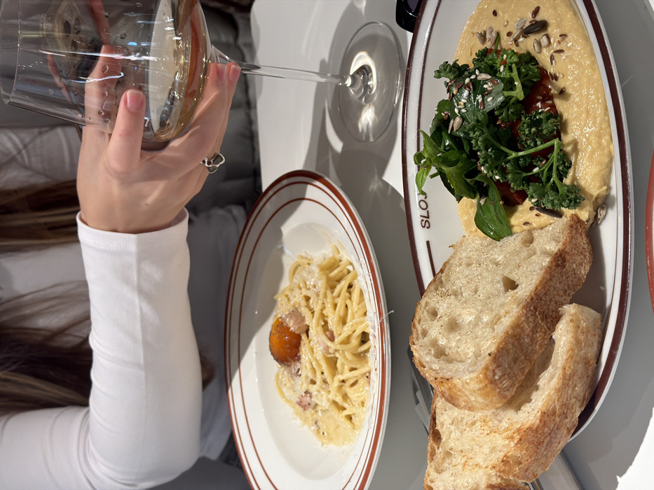 This image depicts an indoor dining scene at Sloja restaurant, located at Sremska 9, Belgrade, Serbia. A person is visible from the chest up, wearing a white long-sleeved top, with long light brown hair. Their right hand, adorned with a silver heart-shaped ring on the ring finger, is holding a clear stemmed wine glass containing a light-colored liquid, likely white wine.
On the white table surface, two primary dishes are presented. In the background, a white bowl with a brown rim holds a serving of spaghetti, topped with a raw egg yolk, grated cheese, and pieces of cured meat (e.g., guanciale). In the foreground, an oval white plate, also with a brown rim and labeled "SLOJA," features a creamy, light-yellow dip, consistent with hummus. This dip is garnished with a variety of green herbs and leaves (e.g., parsley, kale, basil), sun-dried tomato pieces, and a scattering of seeds including sesame, sunflower, and flax seeds. Two slices of rustic, crusty bread are placed on the left side of this plate. A metallic utensil is partially visible beneath the pasta bowl. The lighting is bright, creating reflections on the table and wine glass.