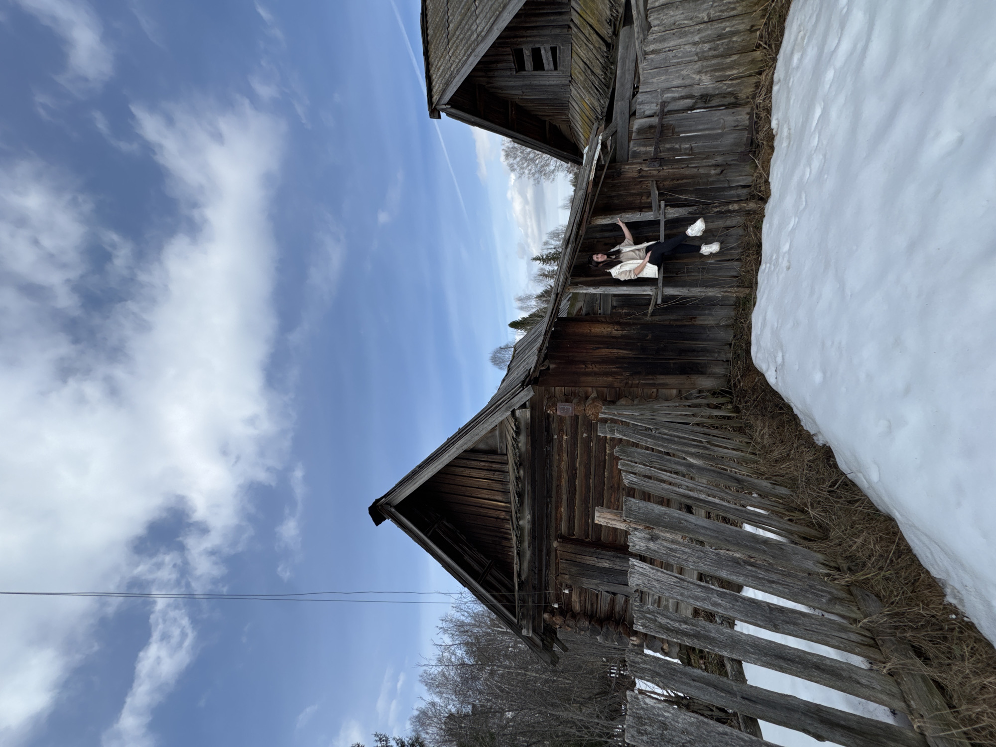 The image depicts a young woman sitting on the dilapidated wooden structure of a building in Вассята, Russia. Snow is visible adjacent to the building. The sky is blue with scattered clouds. The building appears to be old and in disrepair.