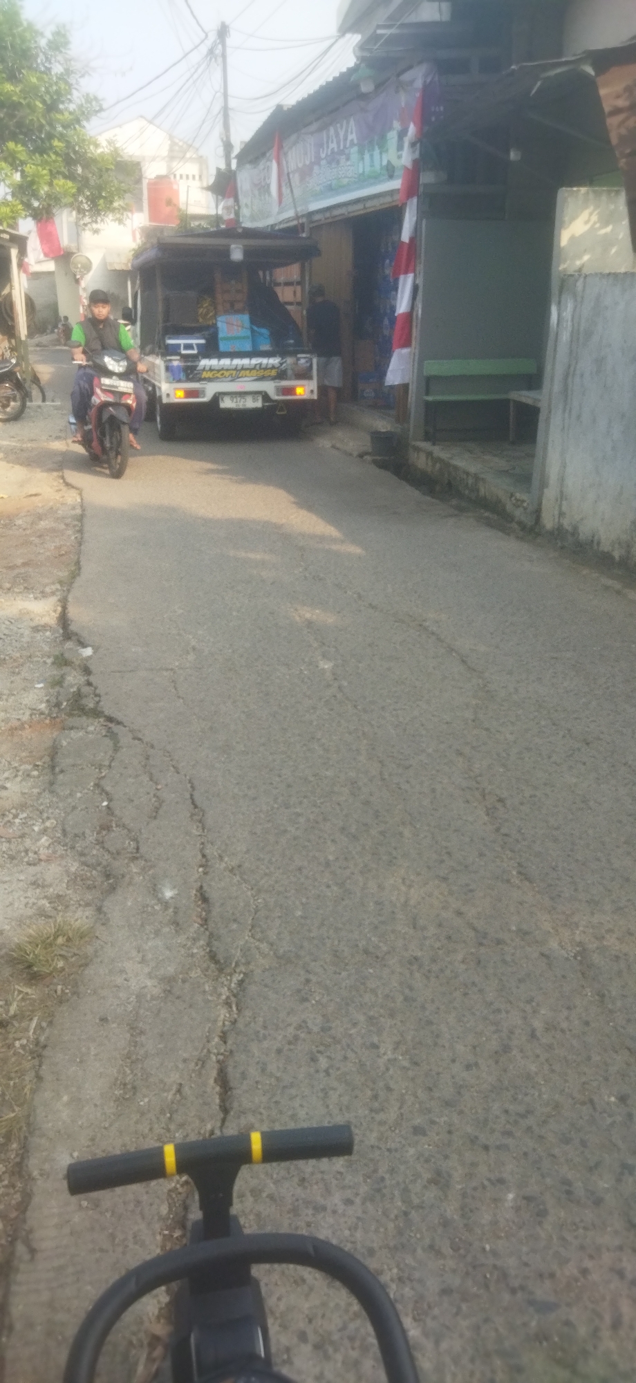 The image depicts a narrow, cracked asphalt road in a densely populated urban area of Depok, Indonesia, possibly a residential alleyway or a service road. A man wearing a green shirt and a cap rides a red motorcycle on the left side of the road. Behind him, a small white utility truck with a canopy is parked, loaded with blue boxes and other items. A banner above a storefront on the right reads "MUJI JAYA" and features red and white Indonesian flags, indicating a shop or business. The environment is characterized by overhead electrical wires, some vegetation, and a simple concrete wall. The foreground of the image shows a black stroller or toy vehicle with yellow accents. The overall scene suggests a typical day in a bustling Indonesian neighborhood.