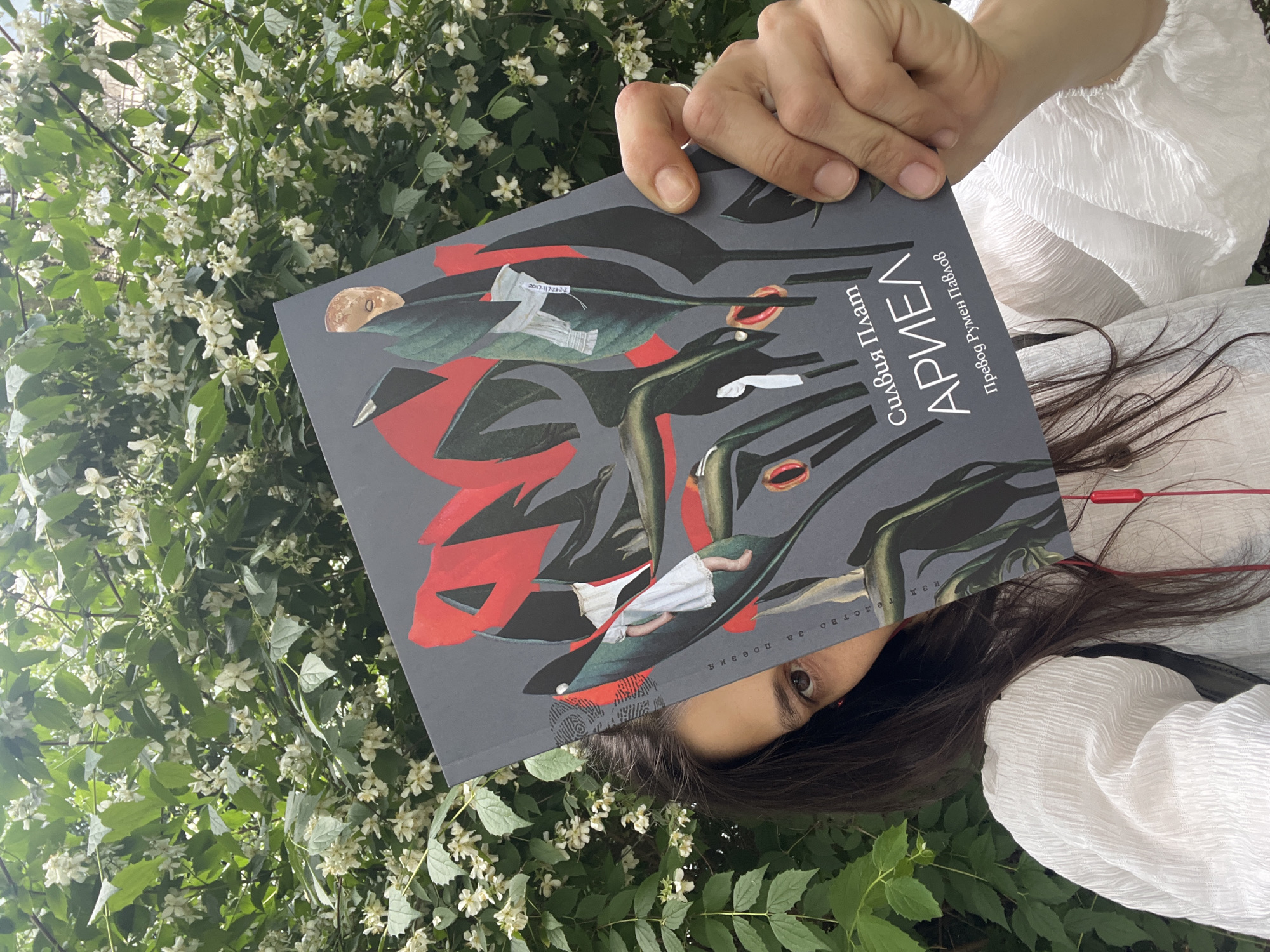 A woman is holding a book in front of her face, partially obscuring her features, against a backdrop of flowering bushes. She is wearing a white textured blouse and red earbuds are visible. The book has a dark gray cover with a surreal illustration featuring red and green abstract shapes, a small white figure in a dress, and a golden mask-like element. Text on the cover reads "Силвия Плат АРИЕЛ Превод Румен Павлов," which translates to "Sylvia Plath ARIEL Translation Rumen Pavlov." The flowering bushes are dense with small white blossoms and green leaves, suggesting an outdoor, possibly garden or park, setting. The overall lighting indicates it is daytime. The scene is captured from a medium close-up angle.