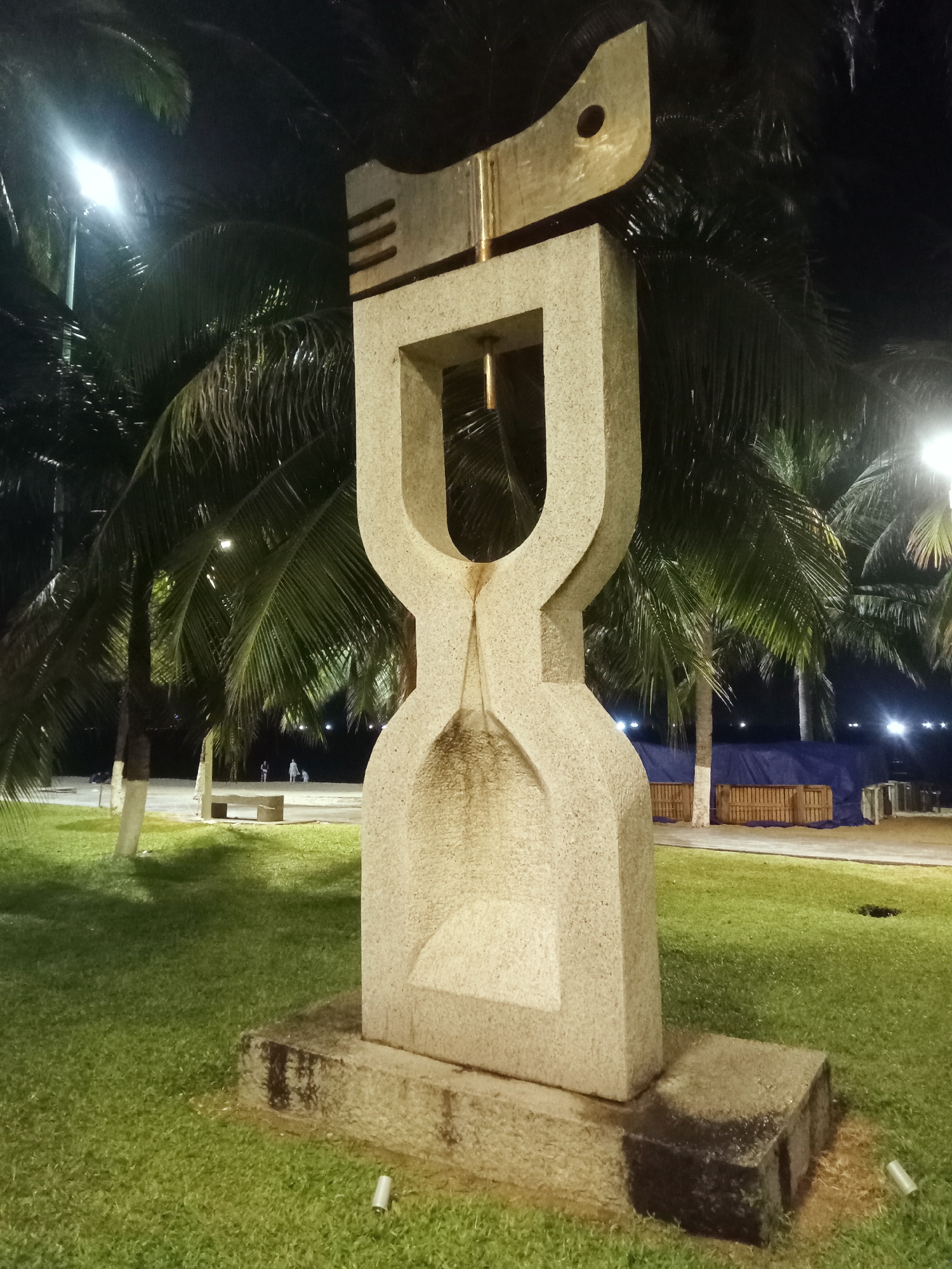 The image shows a sculpture at night in Nha Trang Ward, Vietnam. The sculpture is made of light-colored stone and is shaped like an hourglass with a metal piece on top resembling a fish. The sculpture stands on a small stone base surrounded by green grass. Palm trees are visible in the background, lit by streetlights. In the distance, there are people walking on a paved area. A blue tarp and wooden crates are also visible in the background.