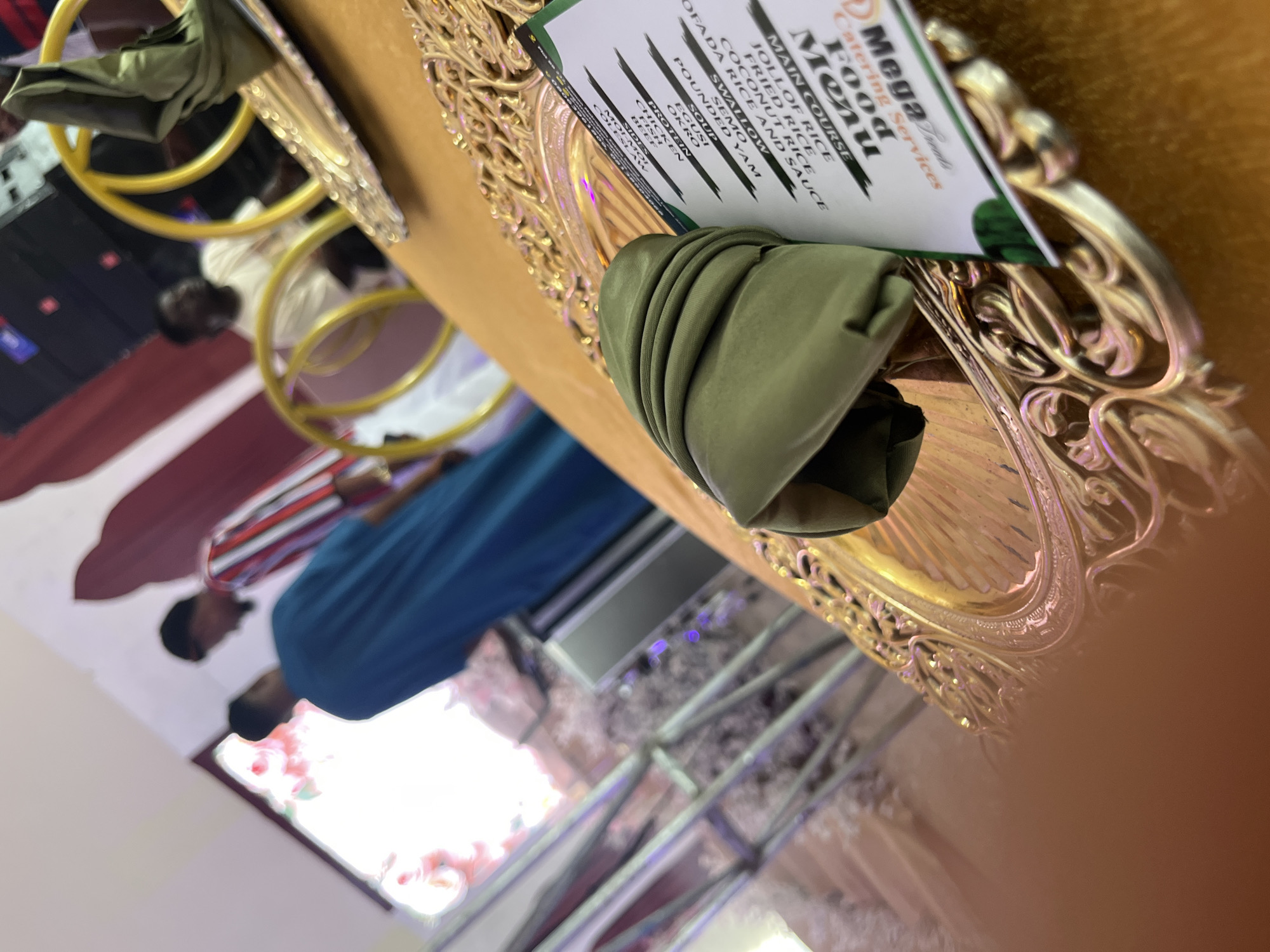 A high-angle, medium shot captures a close-up of a gold ornate charger plate with a folded green napkin on top. The charger is placed on a golden-yellow tablecloth. A menu for "Mega Food Catering Services" is partially visible to the right of the napkin, listing "Main Course," "Fried Rice," "Jollof Rice," "Coconut Rice," and "OFADA RICE AND SAUCE." To the left of the charger, a blurry figure in a blue garment stands with their back to the camera. In the background, several other individuals are present, also out of focus. One person wears a striped shirt. The setting appears to be an indoor event, possibly a banquet or a party, with decorative elements and lighting visible. The overall scene suggests a catered event.