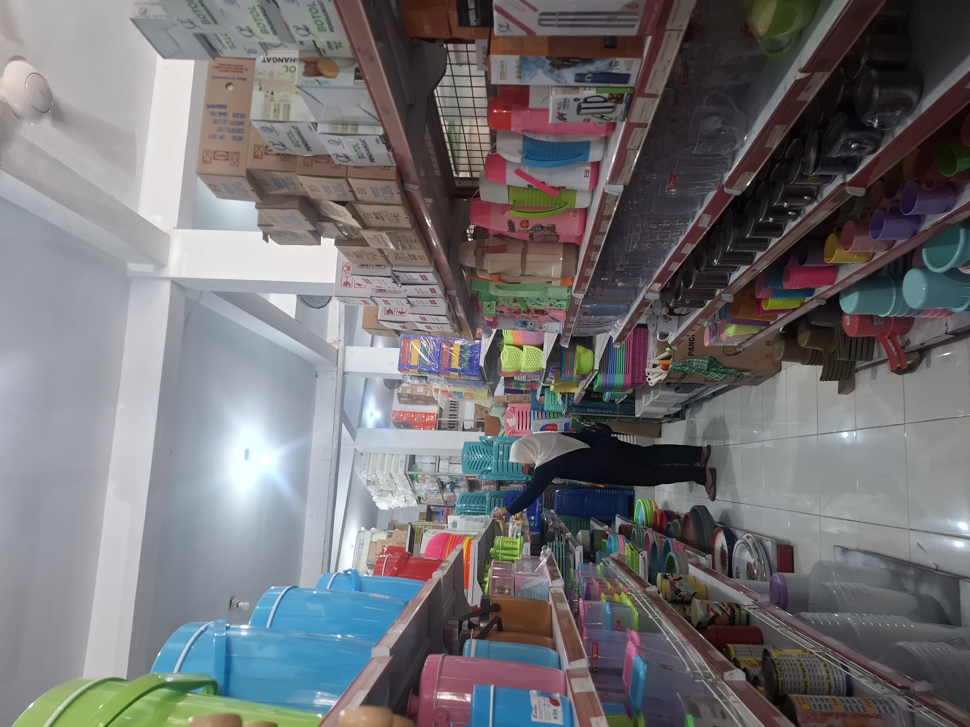 The image depicts the interior of a retail store in Sumberwudi, Indonesia, specializing in household goods. A woman wearing a hijab and a dark-colored outfit is seen browsing the shelves, reaching for an item. The store is brightly lit, with fluorescent lights on the ceiling. Shelves are filled with a variety of plastic containers, buckets, thermoses, and other kitchenware in a range of colors, including blue, green, pink, and red. Boxes are stacked on higher shelves, some labeled with text in Indonesian. A ceiling fan is visible near the top of the frame. The floor is tiled in a light color. The overall impression is one of a well-stocked, organized store.