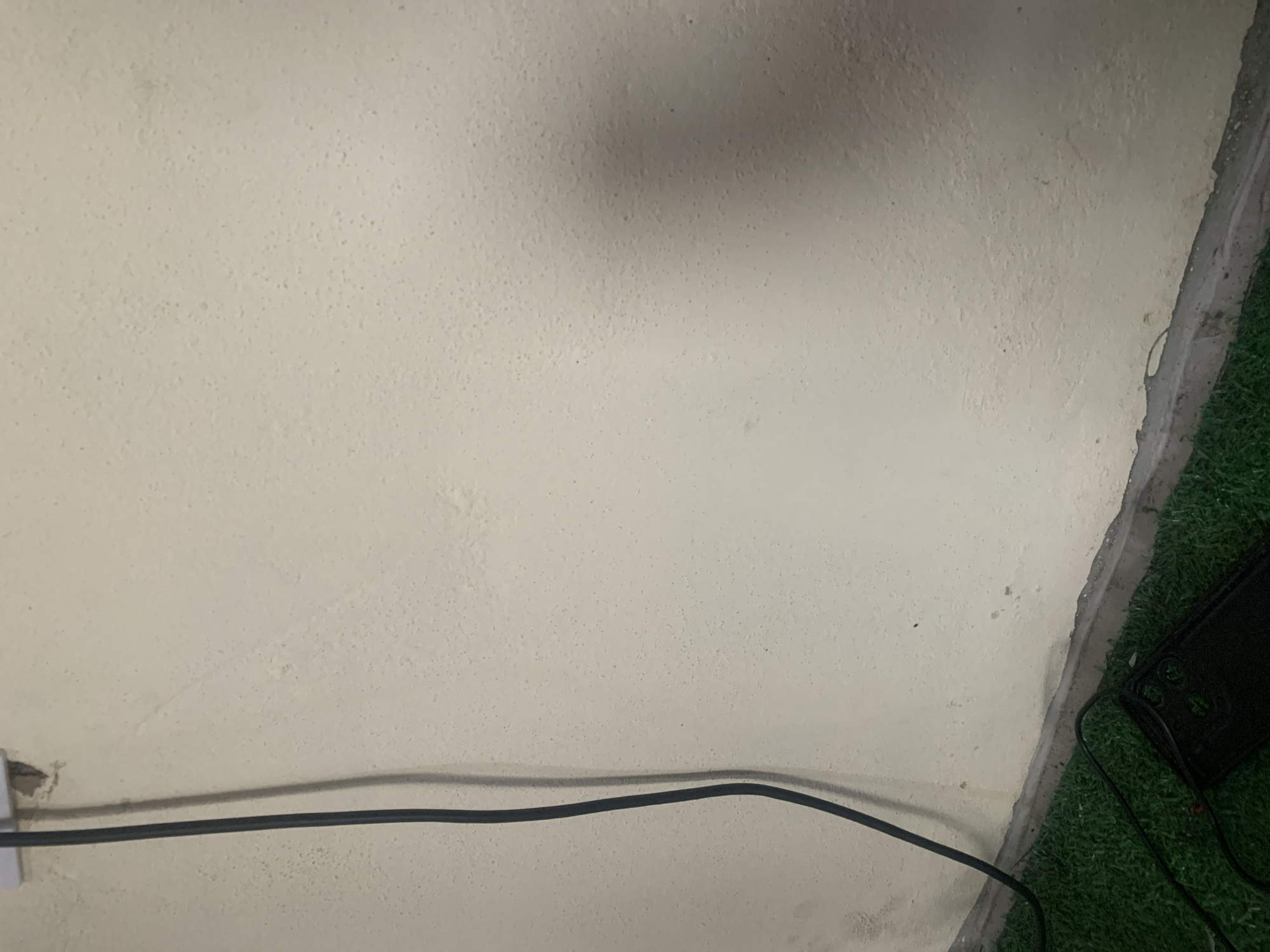 A close-up, eye-level shot captures a section of a textured, light-colored wall in Jauro Yinu, Nigeria. A black electrical cable snakes down the left side of the frame, originating from a white electrical socket near the top edge, which appears slightly damaged. The wall's surface is uneven, with numerous small bumps and irregularities, and subtle shadows suggest it is illuminated by a light source from the right. The lower portion of the image shows a strip of artificial green turf, indicative of an indoor setting or a protected outdoor area. A dark, rectangular object, possibly an electronic device or a mat, rests on the turf near the bottom left corner, with another black cable extending from it. The scene is devoid of any people or discernible action, presenting a still, mundane environment.