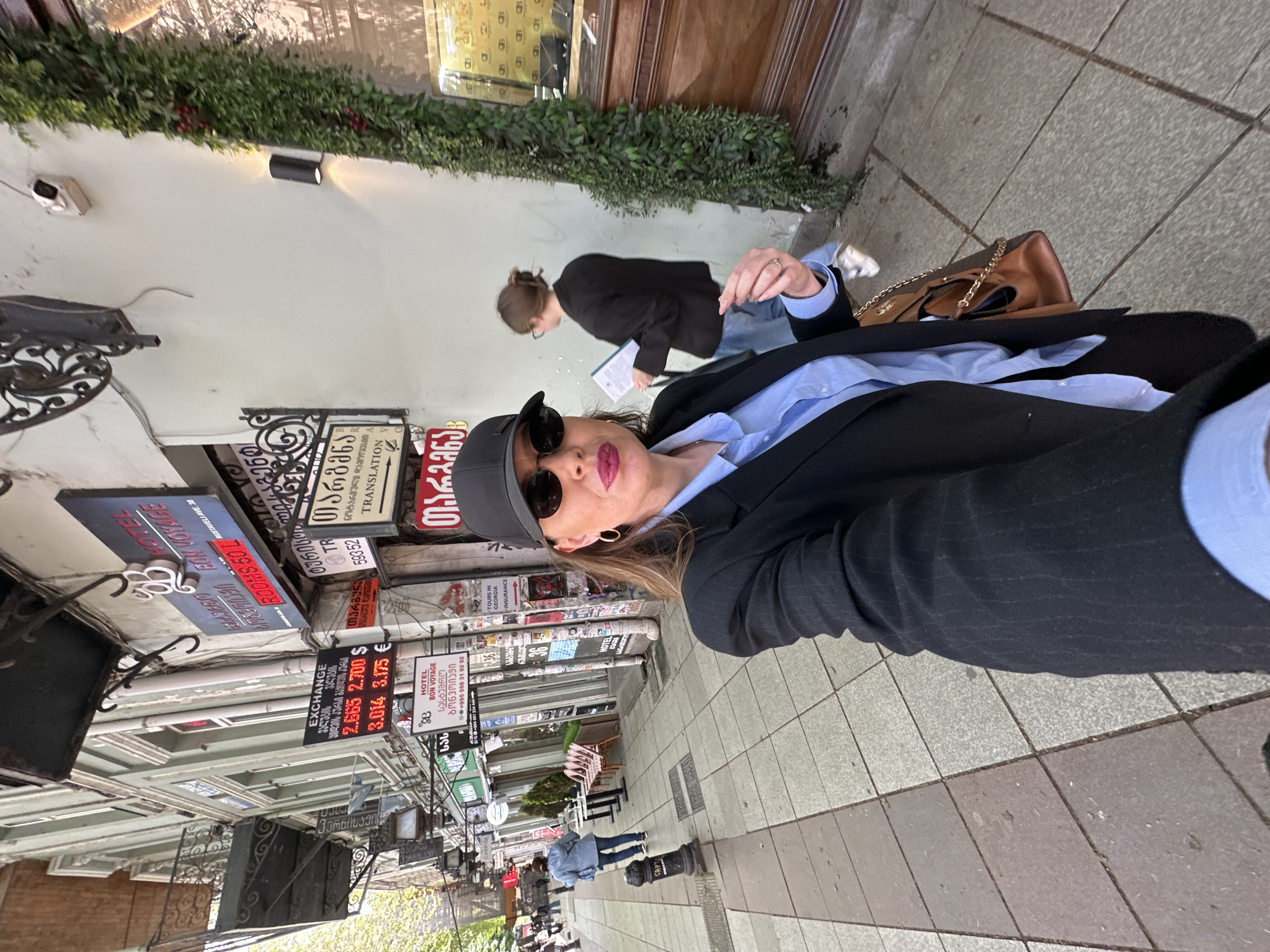 A woman in a dark blazer, light blue shirt, sunglasses, and a baseball cap stands on a sidewalk in Tbilisi, Georgia. Another person is behind her, holding a clipboard and wearing a black blazer and jeans. Several signs in Georgian and English hang from buildings, advertising hotels, translation services, and currency exchange rates. A brown handbag with gold chains sits on the ground next to the woman in the blazer. Other people can be seen walking further down the sidewalk. The environment consists of a sidewalk with gray tiles and buildings decorated with greenery. The currency exchange rate is $2.665 / $2.700. The hotel is Hotel Bon Voyage.