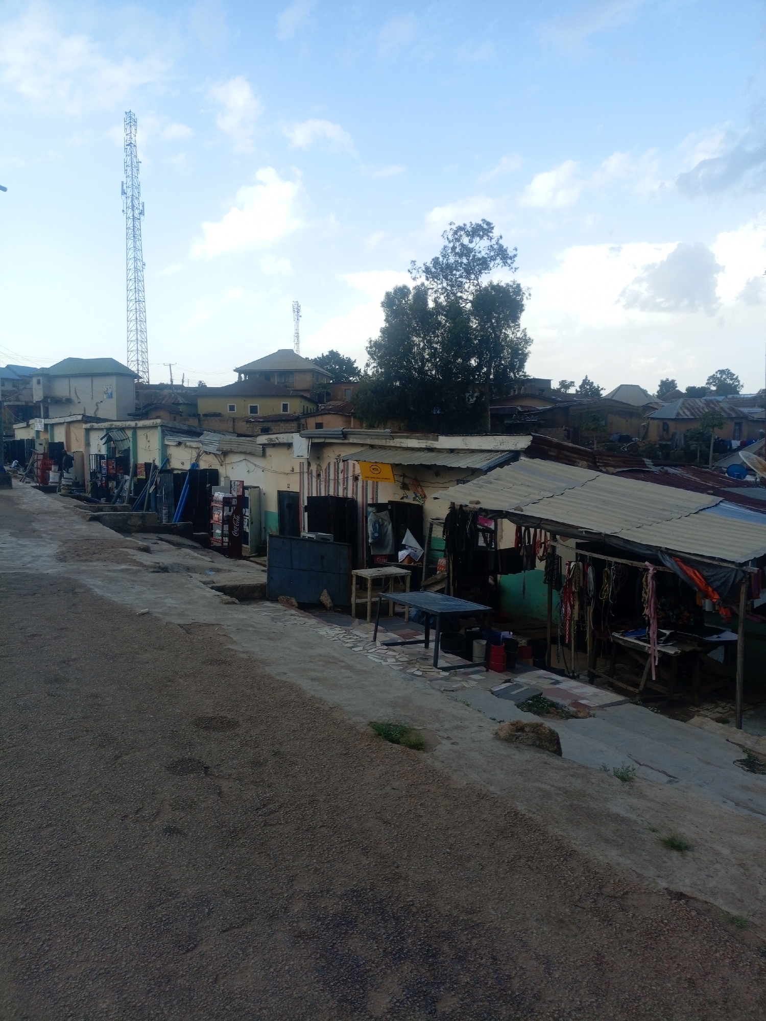 The image depicts a street scene in Jos, Nigeria, likely a commercial area or market. A paved road occupies the foreground, curving to the left and sloping upwards. The background is filled with a row of small shops and buildings with corrugated metal roofs, some appearing weathered. Several of these stalls display various goods, including what appear to be clothing items, textiles, and possibly tools or hardware. In the middle ground, a Coca-Cola refrigerator is visible outside one of the shops. Further back, more buildings of varying heights and construction styles are present, with some showing signs of disrepair. A tall telecommunications tower stands prominently in the distance, and another, shorter one is visible to the right of it. Trees with dense foliage are scattered throughout the scene, some taller than the surrounding structures. The sky is partly cloudy, with patches of blue and diffused light suggesting it may be late afternoon or early morning. No people are clearly visible in the image.