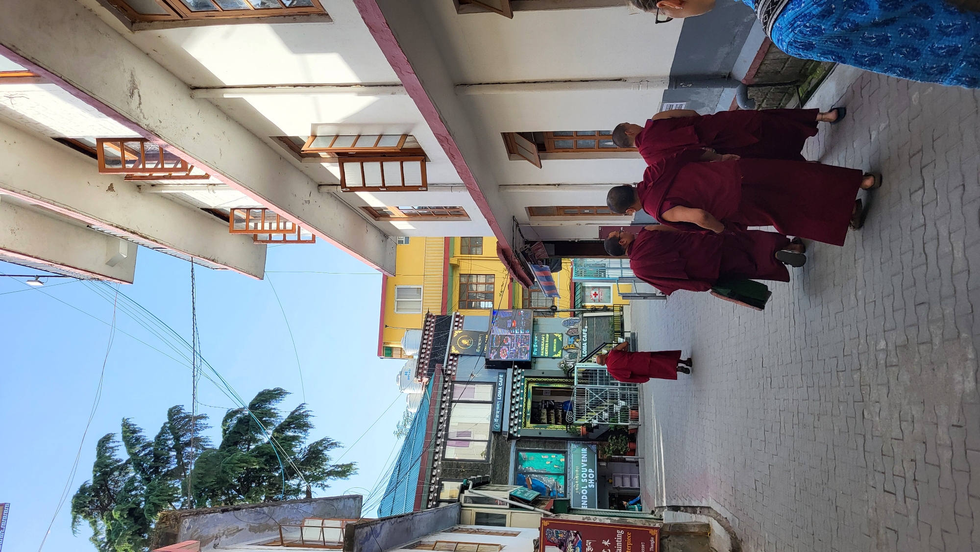 The image captures a street scene in Dharamshala, India, likely a commercial area near a temple or monastery. A group of Buddhist monks clad in maroon robes walks along a paved walkway. Street-level shops line the walkway and are marked with signs, including one for "ENDOL SOUVENIR SHOP." The buildings exhibit a mix of architectural styles and colors. Overhead electrical wires crisscross the sky, and trees are visible in the background. The lighting suggests a sunny day.