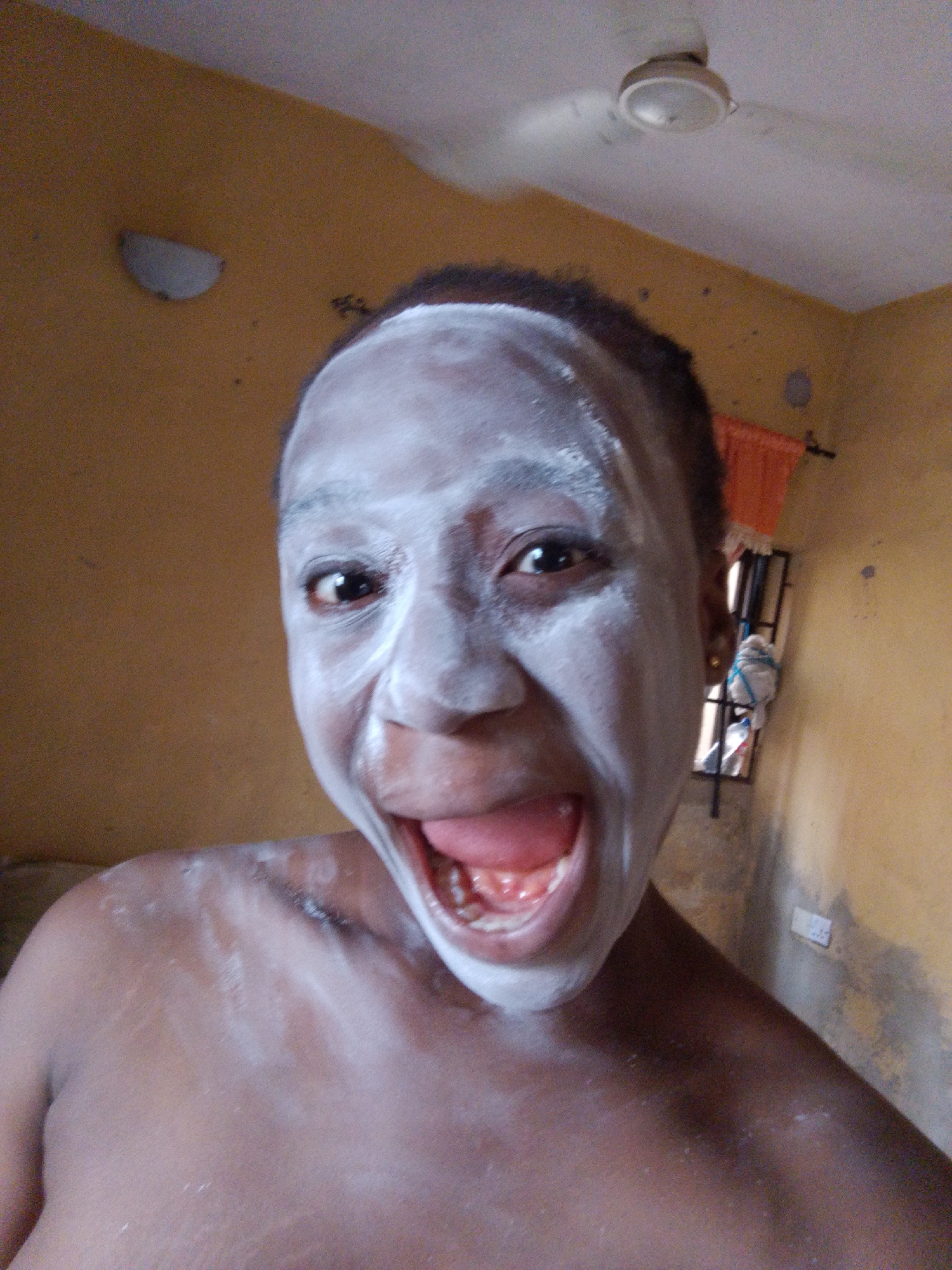 A close-up shot depicts a person with dark skin, gender unclear, face covered in white powder, mouth open, and teeth visible. They are indoors. The walls are painted a dull yellow. In the background, there's a window with a curtain and a ceiling fan. Location is not discernible to Umuahia, Nigeria, or any other specific location.