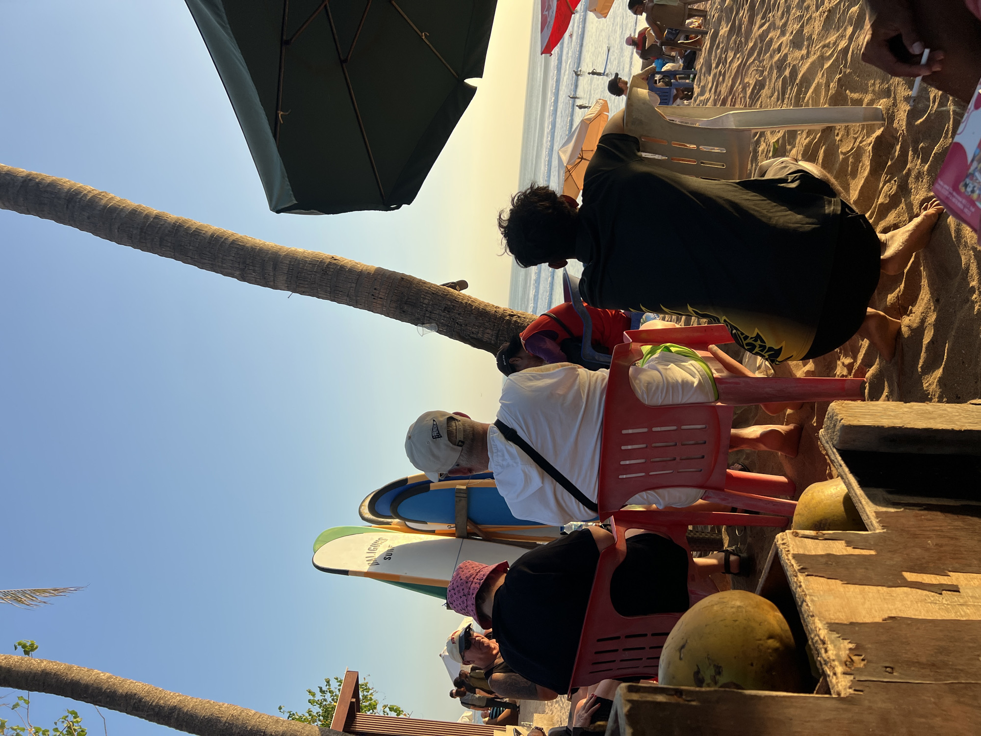 In Kuta, Indonesia, an outdoor cafe on a sandy beach is depicted at sunset. A cluster of palm trees dominates the foreground, with one large tree trunk diagonally bisecting the frame, casting a shadow over the scene. In the middle ground, several people are seated on red and white plastic chairs, facing the ocean. Two individuals in the foreground are seen from behind, one wearing a white hoodie and the other a black t-shirt. Behind them, surfboards are propped up against a wooden structure. Further back, more people are visible, some standing and some sitting at tables under umbrellas. The ocean stretches to the horizon, with gentle waves and a few individuals in the water. The sky is a clear, pale blue, transitioning to a warm orange hue near the horizon, indicative of sunset. In the immediate foreground, a rustic wooden structure holds two coconuts. The overall atmosphere suggests a relaxed evening at a beachside establishment.