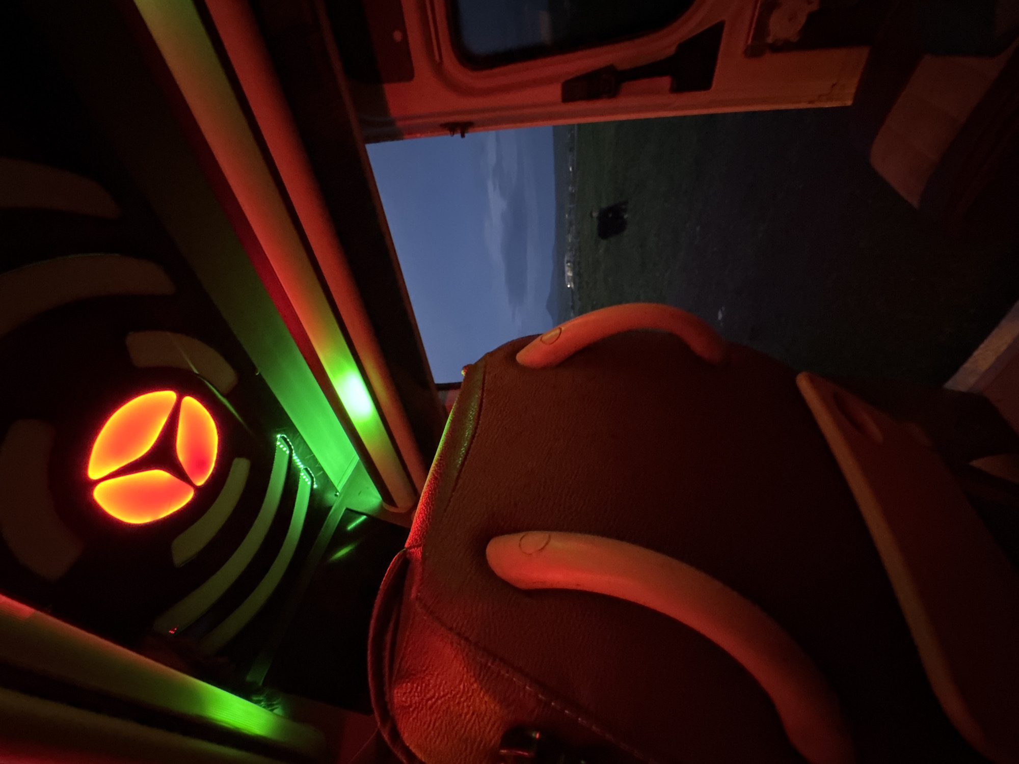 The image depicts the interior of a vehicle, likely a van or bus, with customized lighting. A circular orange light fixture with a three-lobed design is mounted on the wall. Green LED strip lights are visible as well.
A vehicle seat with a reddish-brown leather or vinyl covering is in the foreground. Two light-colored plastic or composite grab handles are attached to the seat.
Through the open rear doors of the vehicle, a twilight or nighttime landscape is visible. A dark, indistinct shape, possibly an animal, is visible in the dark field. Distant lights, suggesting a settlement, are visible on the horizon. The city of Anipemza, Armenia is not identifiable in the image.