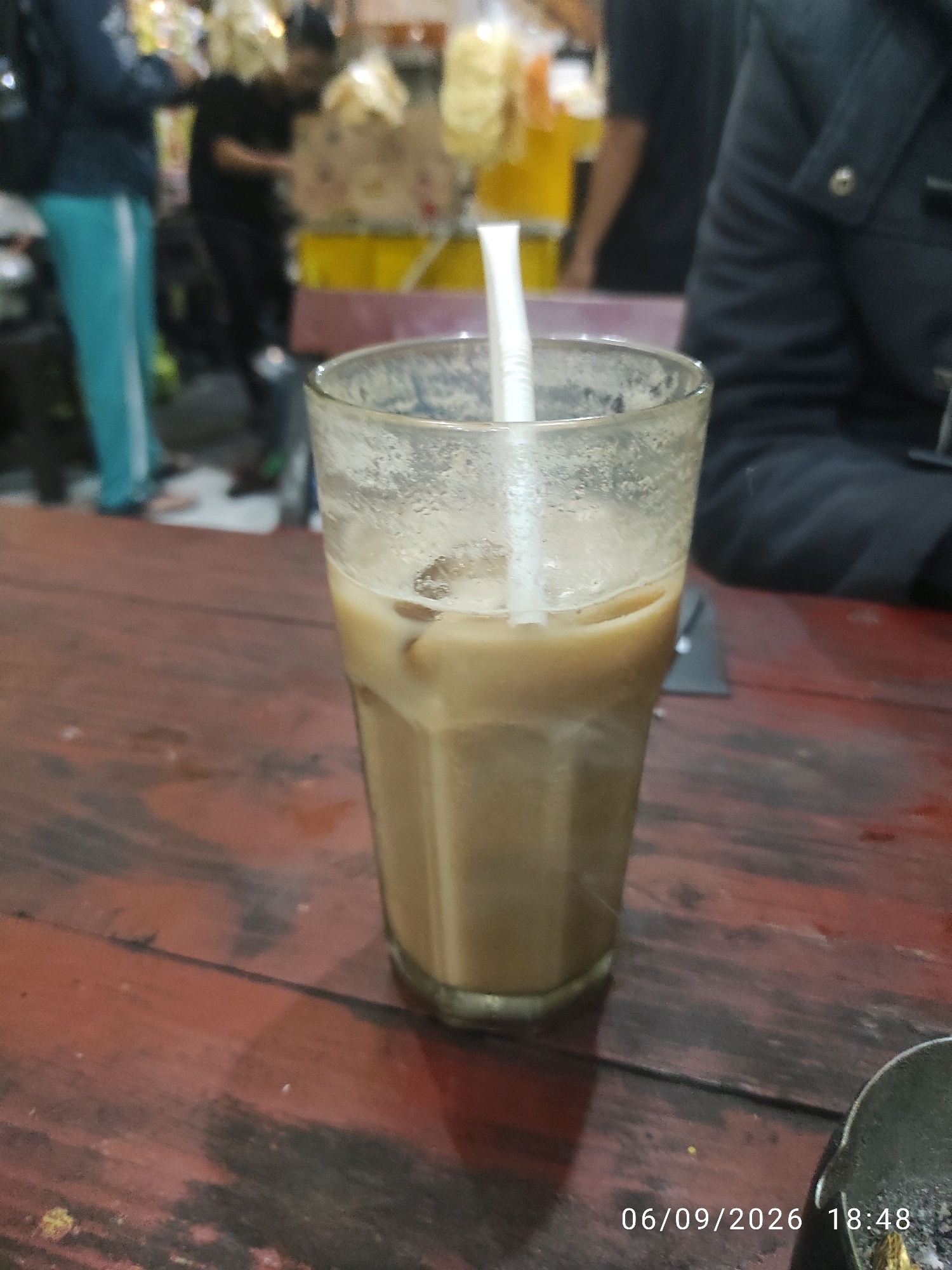 A close-up shot focuses on a glass filled with iced coffee, placed on a weathered, reddish-brown wooden table. A white straw extends from the liquid. In the blurred background, several individuals are visible, along with stalls displaying food items. The overall setting appears to be an outdoor market or food stall area in Surabaya, Indonesia. The timestamp "06/09/2026 18:48" is superimposed on the lower right of the image.