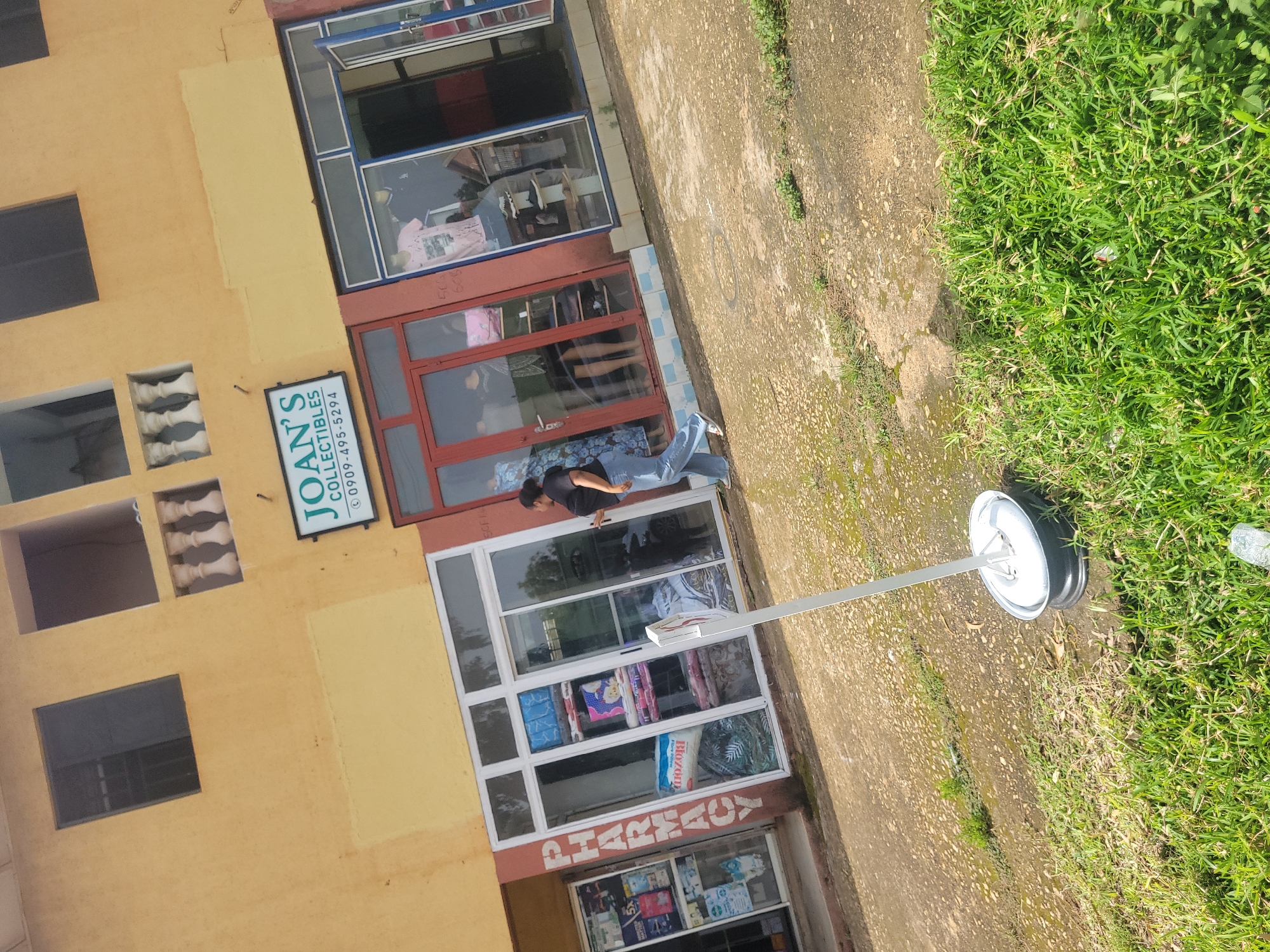 The image shows a storefront in Jos, Nigeria, with the name "JOAN'S COLLECTIBLES" displayed above a doorway. The building is painted yellow with brown trim. To the left of "JOAN'S COLLECTIBLES" is a sign that reads "PHARMACY". A woman in blue jeans and a dark top is walking away from the storefront on a paved area. The ground is covered in patches of grass and has a mottled, wet appearance. A white pole with a silver base stands near the foreground. In the background, there are windows and balconies on the upper level of the building.
