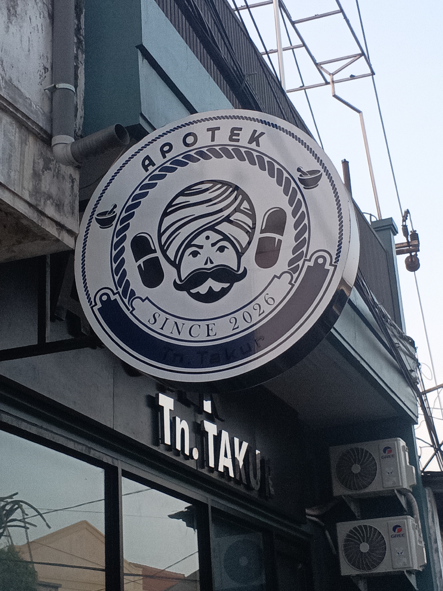 The image captures a close-up view of the signage for "Apotek Tn. Tayu Ku," an establishment in Surabaya, Indonesia. The primary focus is a large, circular sign with intricate detailing. The sign features the word "APOTEK" arching above a stylized illustration of a person with a turban, a mustache, and a bindi on their forehead, flanked by two pills. Below this illustration, the text "SINCE 2026" is prominently displayed within a dark blue banner. Beneath the circular sign, the name "Tn. Tayu Ku" is spelled out in white, three-dimensional lettering against a dark facade. The scene also shows two air conditioning units mounted on the building's exterior wall, one above the other. The overall setting appears to be an urban environment, with glimpses of electrical wiring and structural elements of a building visible in the background.