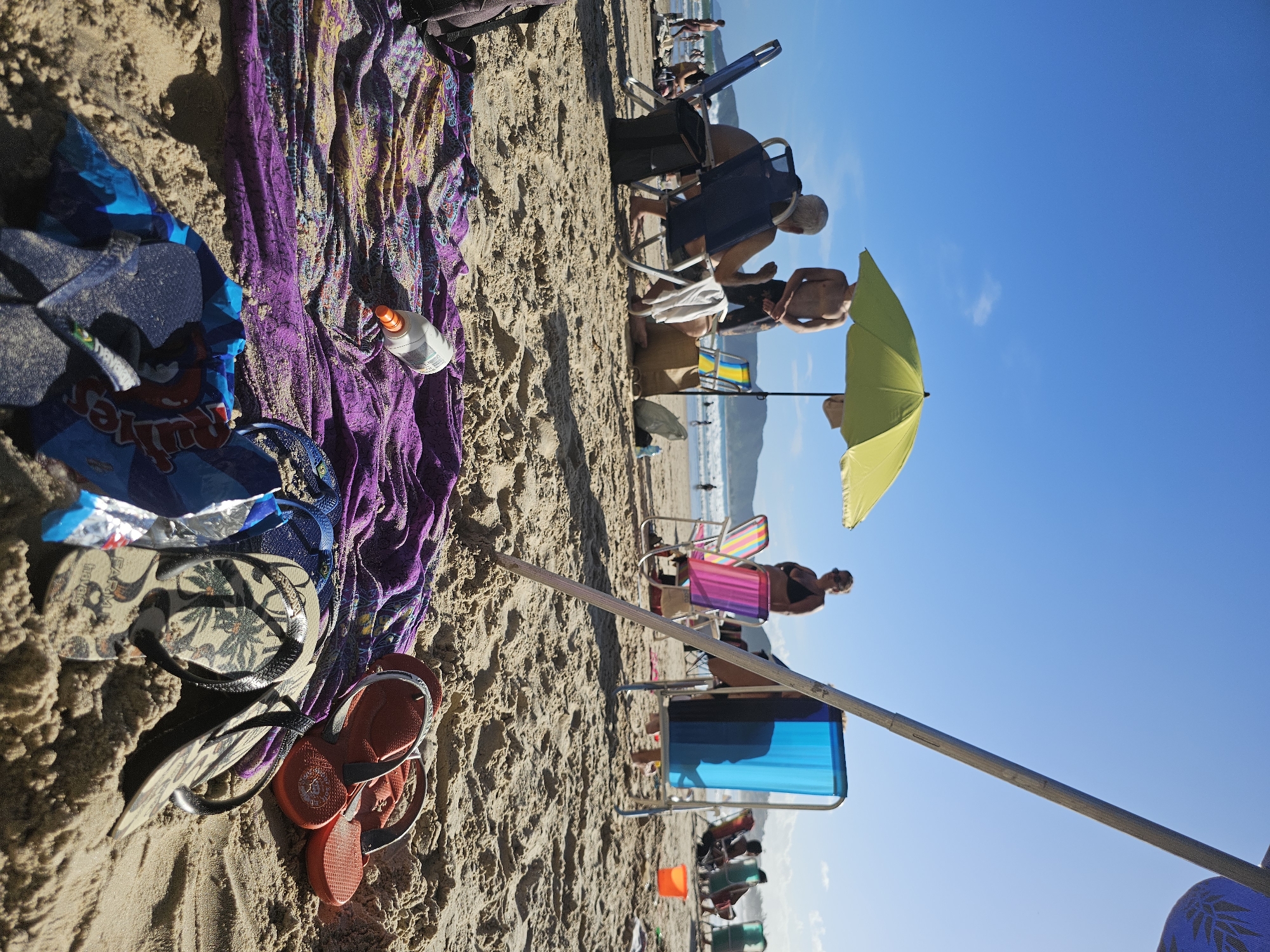 The image depicts a beach scene in Armação Dos Búzios, Brazil. In the foreground, there are beach items such as a purple towel, a bag of chips, sunscreen, and two pairs of flip-flops (one pair is red). Further into the scene, individuals are visible on the beach. Some are sitting in beach chairs under a yellow umbrella, while others are standing. In the background, the ocean is visible. The sky is blue.