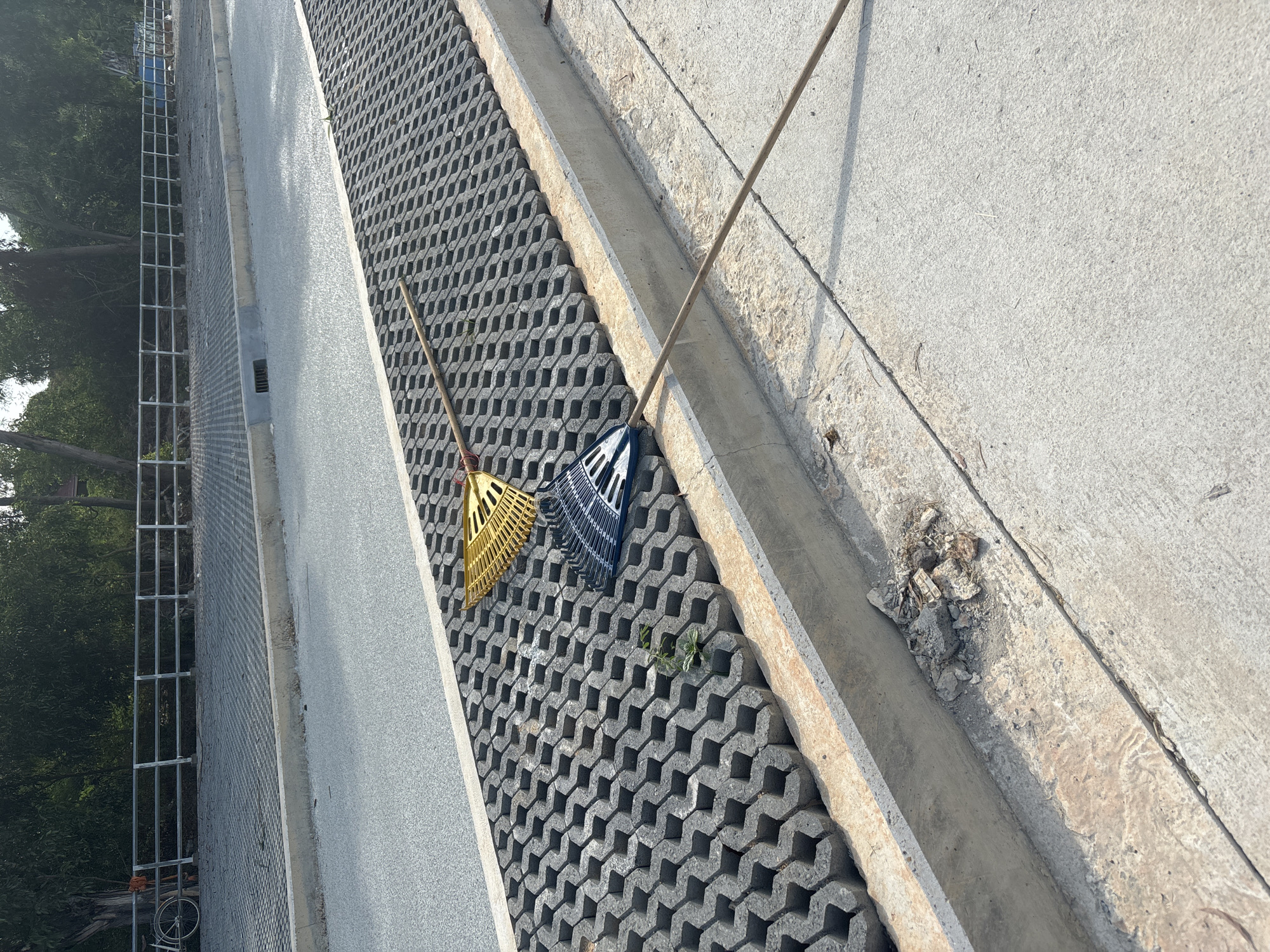 The image depicts a street scene in บ้านบางรักษ์, Thailand. The view is from an elevated perspective, looking down at a section of sidewalk and permeable paving. Two leaf rakes, one yellow and one blue, lie discarded on the permeable paving. A broom also rests nearby. Adjacent to the permeable paving is a concrete drainage ditch containing debris. A newly paved gray sidewalk borders the permeable paving on one side. In the background, a metal fence is visible, with trees and a partial view of a building beyond. A bicycle is partially visible in the lower left corner. The scene appears to be during daylight hours under bright sunlight.