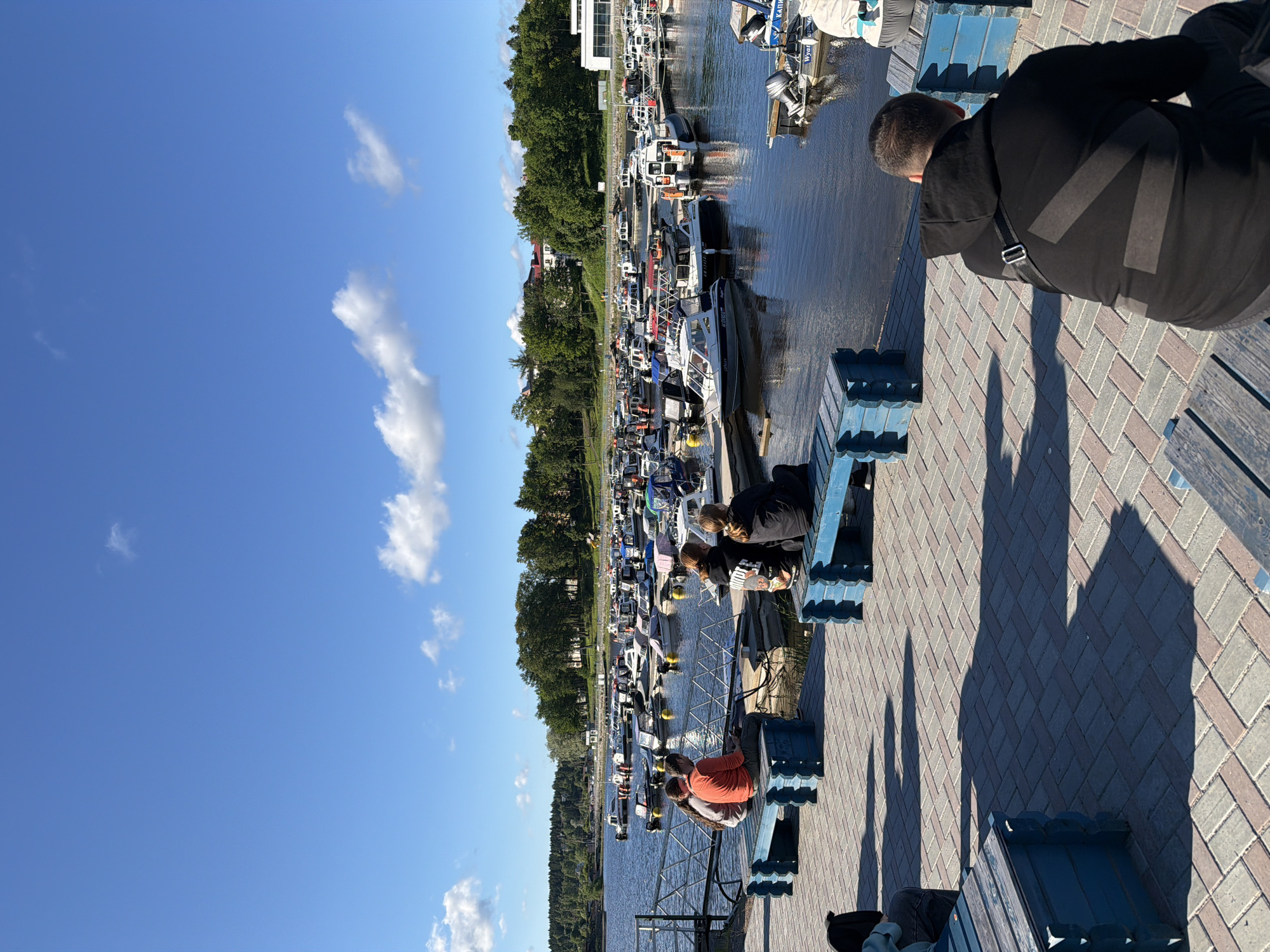 The image captures a sunny day at a marina in Sortavala, Russia. A body of water, likely a bay or lake, is filled with numerous moored boats of various sizes, ranging from small motorboats to larger yachts. The boats are docked along wooden piers, and yellow buoys are visible in the water.
Along the waterfront promenade, paved with brickwork in a herringbone pattern, several blue wooden benches are placed. On one of these benches, three people are seated, with two in the foreground facing away from the camera and one in the background. A fourth person is seated on another bench in the foreground, also facing away. Further down the promenade, another group of three people are seated on a blue bench, facing the water.
Behind the marina, a lush green treeline stretches along the shoreline, with buildings visible among the trees. The sky is a bright, clear blue with a few scattered white clouds. The sunlight casts distinct shadows on the promenade, indicating a bright and clear day. The overall scene depicts a leisurely atmosphere at a waterside recreational area.