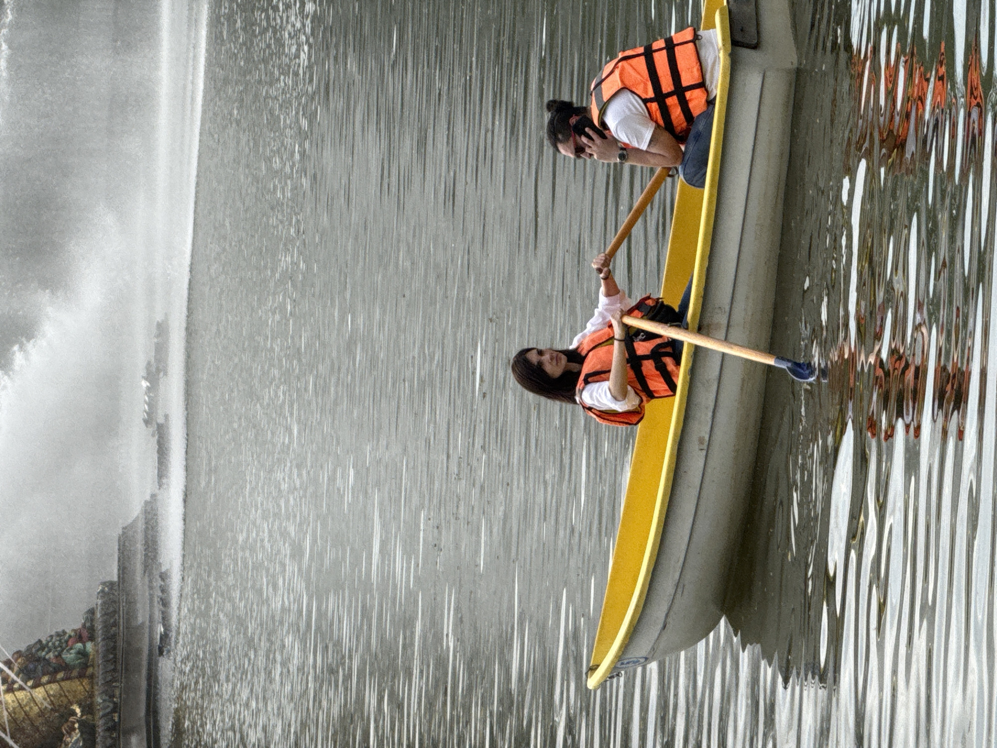 Two individuals are in a yellow and grey boat on a body of water. Both are wearing orange life vests. One person is seated and appears to be rowing the boat, while the other is standing and seems to be looking at something in the distance. The boat is near a structure with a cascading waterfall. The location appears to be in Moscow, Russia.