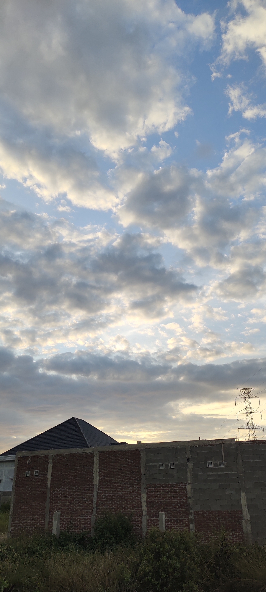 This image depicts an overcast sky with scattered clouds, possibly at dusk or dawn, over an urban or semi-urban landscape. In the foreground, there is a partially constructed brick wall with concrete pillars. Behind the wall, a dark-tiled pyramidal roof of a house is visible. To the right of the house, a tall metal electricity pylon stands against the sky. The lower portion of the image is filled with dry grass and green bushes. The scene is devoid of any people or visible actions. Based on the provided information, the image is located in the city of Lhokseumawe, Indonesia.