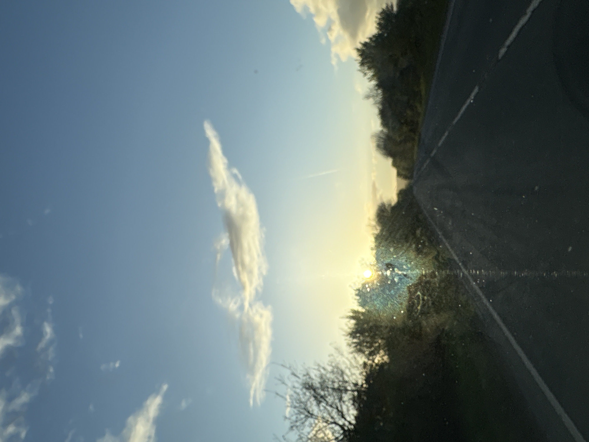 The image depicts a view from inside a vehicle, possibly a car, looking out onto a rural scene. The sky is blue with some scattered white clouds and a contrail from an airplane. The sun is visible near the horizon, obscured by trees. A road with a white line on it is seen on the right side of the image. The location is near Oswestry, United Kingdom, based on geographical context. No people are visible in the frame.