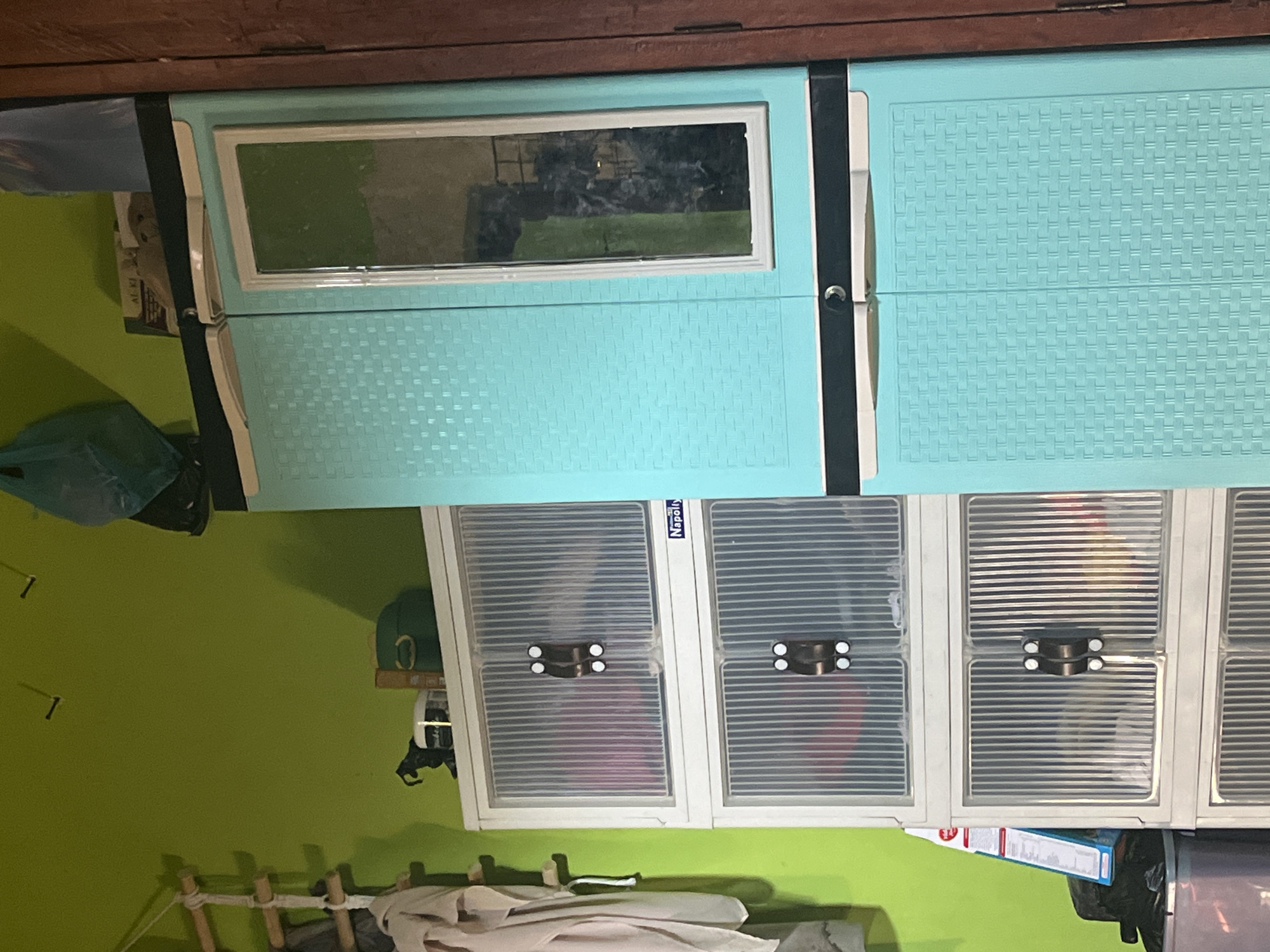 A turquoise plastic wardrobe with a mirrored door stands against a lime green wall. To its left, a stack of white plastic drawers with transparent ribbed doors is visible, containing various items. A wooden ladder with a towel hanging from it is attached to the green wall. On top of the white drawers, several objects are placed, including a black horse figurine and a green container. In the background, a wooden door frame is partially visible. The scene appears to be an indoor storage area in Bumi Mulyo, Indonesia, likely a home or a small business.