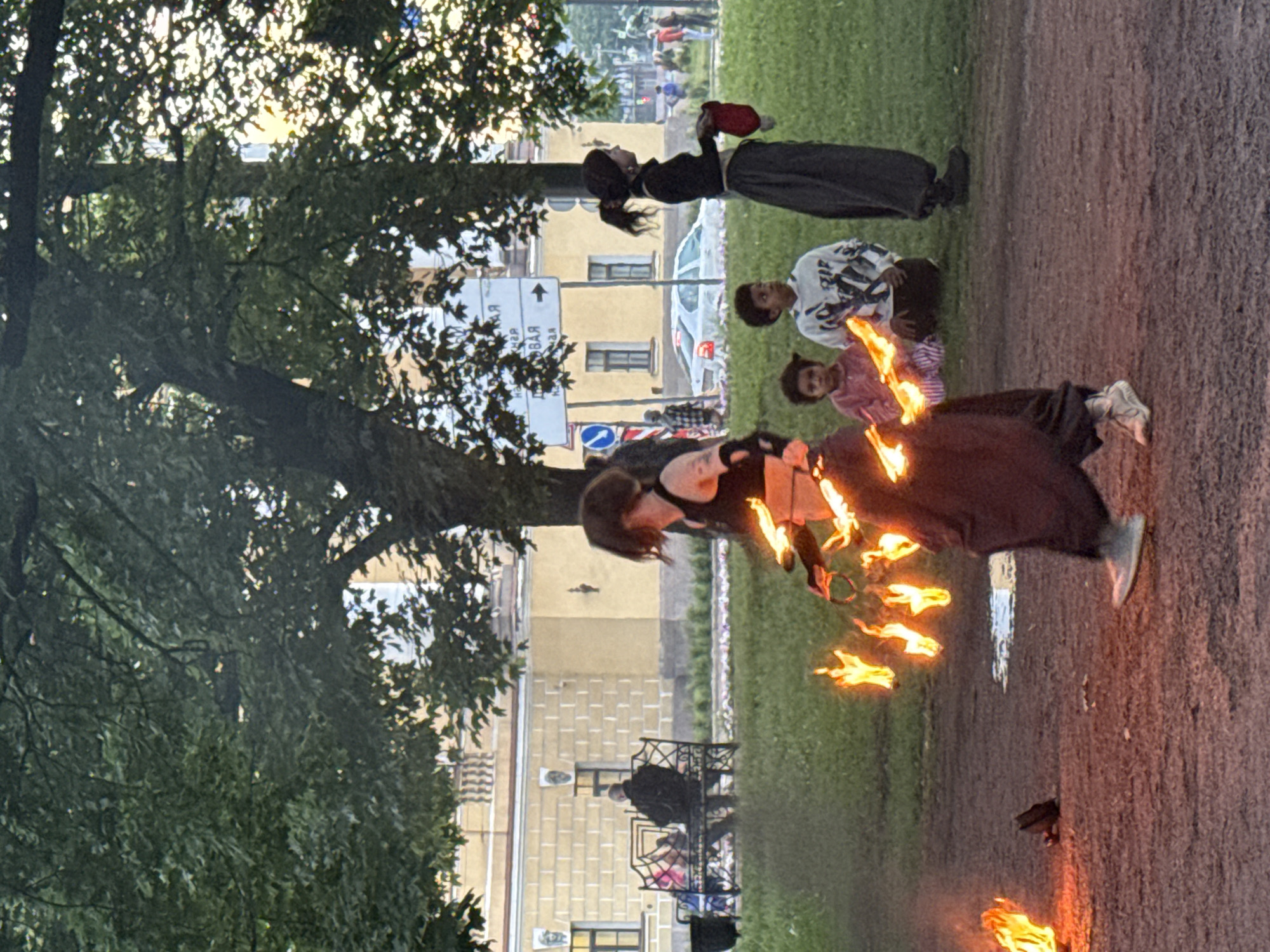 The image depicts a street performance in a park in Saint Petersburg, Russia, likely during daylight hours. In the foreground, a performer is engaged in fire dancing, holding flaming props that trail sparks and light. The performer wears dark, loose-fitting clothing, including a dark top and a long, flowing skirt, with a bracelet on their left arm. They are moving across a dirt path with patches of grass. To the left, another fire is burning on the ground, near a dark, pot-like object.
In the mid-ground, a group of onlookers is scattered across the grass. A woman in a black top and a dark, long skirt stands facing away from the camera, holding a red cloth. Beside her, two children, a boy and a girl, are seated on the grass. The boy wears a white hoodie with a graphic print, and the girl wears a pink jacket. Further back, a park bench is occupied by several individuals, including a man in a dark jacket.
Behind the performers and onlookers, a light yellow building with classical architectural elements, including columns and decorative elements above windows, is visible. Trees with dense green foliage partially obscure the building and the sky. Signs with Cyrillic text and directional arrows are also present. The overall scene suggests a public gathering or event in a park setting within the city.