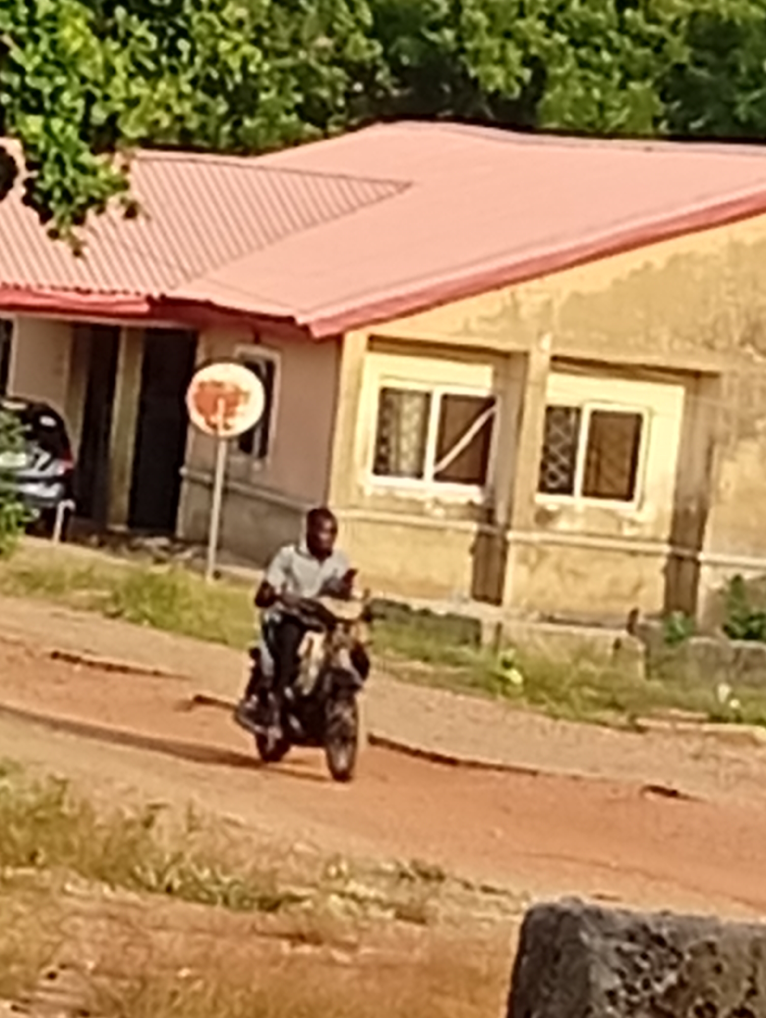 A man is riding a motorcycle on a dirt road in front of a one-story building. The building has a pink roof and tan walls with two windows. There is a car parked in front of the building and a tree in the background. The location is Unknown, Unknown.