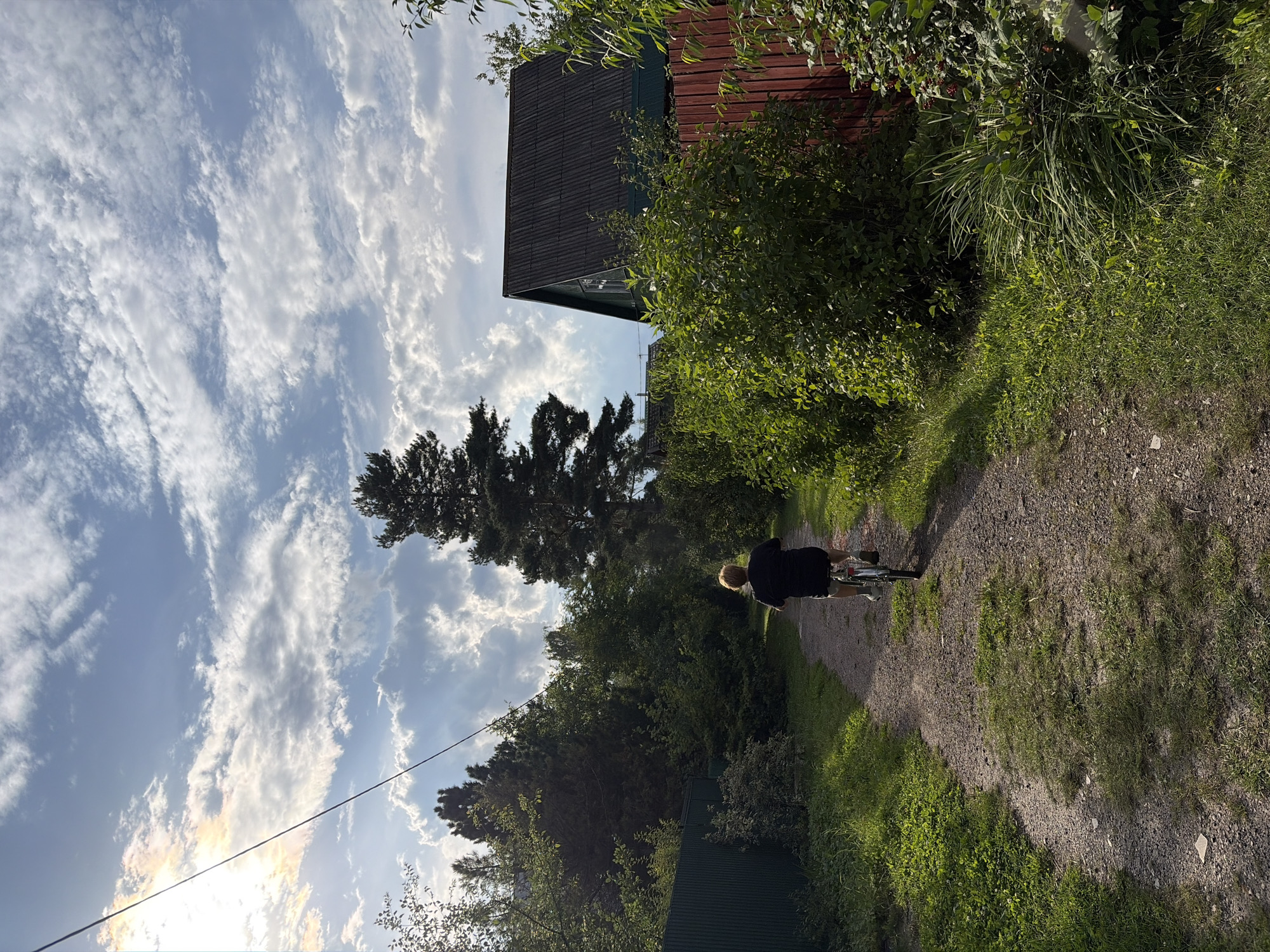 A person rides a bicycle on a gravel path in a rural area, likely the city of Elektrotekhnik SNT, Russia, under a cloudy sky with sunbeams breaking through. The path is bordered by lush green vegetation and trees. On the right side of the path, a dark-colored house with a pitched roof and a red fence are visible. The person on the bicycle is seen from behind, wearing a dark t-shirt and shorts.
