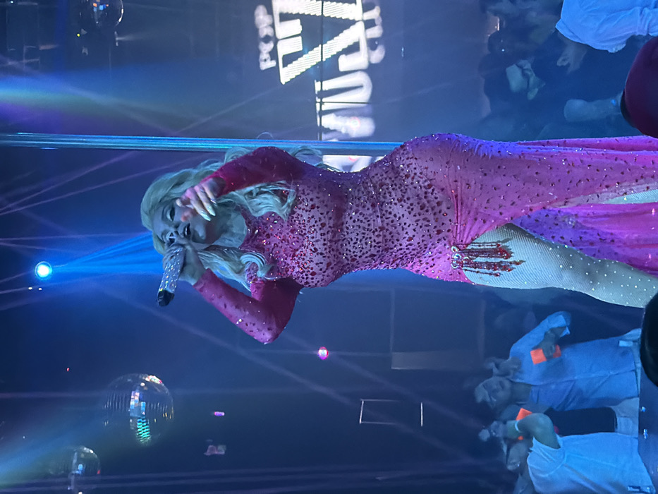 A blonde woman in a pink, long-sleeved, rhinestone-studded dress with a high slit sings into a microphone on stage. The stage is lit with blue spotlights and has disco balls. People are in the background. The words "POP DIVA" are displayed in bright lights above the stage.