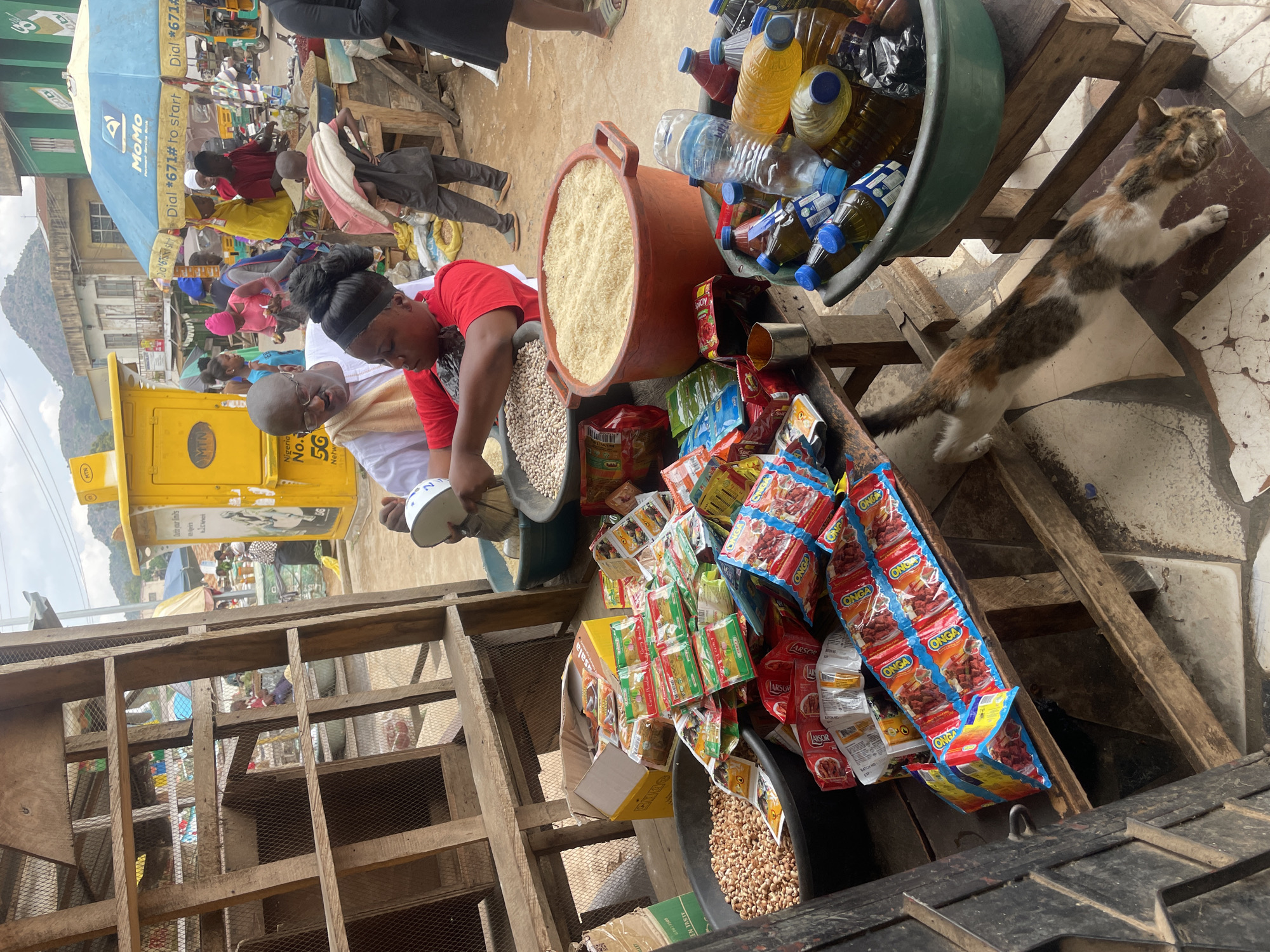 This image depicts an outdoor market scene in Jos, Nigeria, identifiable by the distinctive hilly landscape in the background, the MTN kiosk, and the MoMo Payment Service Bank umbrella.
In the mid-ground, a man in a white tunic and glasses is engaged in a transaction with a woman in a red top. He is holding a white measuring scoop over a black plastic basin filled with light-colored beans. Another black basin to the left contains what appears to be peanuts. Adjacent to these, a large orange pot holds a quantity of white rice. A wooden table in the foreground is covered with numerous small packets of seasonings, including "Curry Powder" and "Onga" brands, some spilling from a yellow cardboard box. To the right, another large plastic basin contains various bottled cooking oils and a plastic water bottle.
The ground is a mix of dirt and patterned tiles. A calico cat with white, brown, and black fur is stretching on the tiled section of the floor in the lower right.
In the background, a prominent yellow MTN kiosk displays "Nigeria No. 1 5G Network" branding. Next to it, a blue umbrella for "MoMo Payment Service Bank" features the text "Dial *671# to start." Beyond the market activity, residential and commercial buildings are visible, with distinct green and brown hills rising in the distance under a clear sky. A young boy carrying a bundle on his back walks through the background market area. Parts of a rustic wooden market stall or structure frame the left side of the foreground.