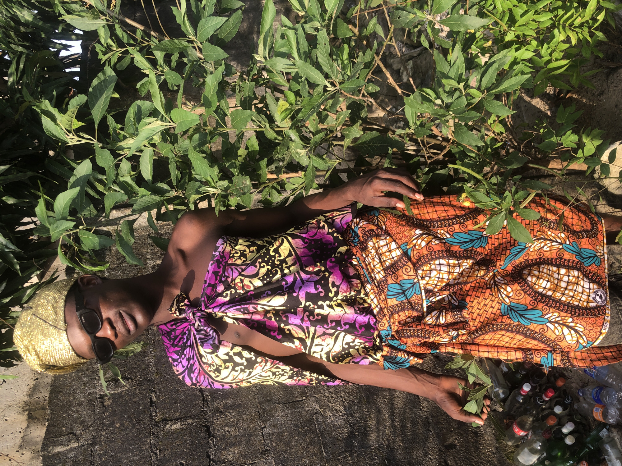 A young man stands outdoors, leaning against a textured, grey stone wall. He is wearing a gold, metallic head wrap, dark sunglasses, and traditional African clothing. The fabric draped over his torso and around his waist features vibrant, intricate patterns in shades of purple, yellow, black, and orange with turquoise accents. To his right, a lush green bush with many leaves grows tall, partially obscuring the wall. In the lower left corner of the image, a collection of discarded plastic and glass bottles lies on the ground. The lighting suggests it is daytime, with sunlight casting shadows. The overall setting appears to be an outdoor urban environment in Ado, Nigeria.