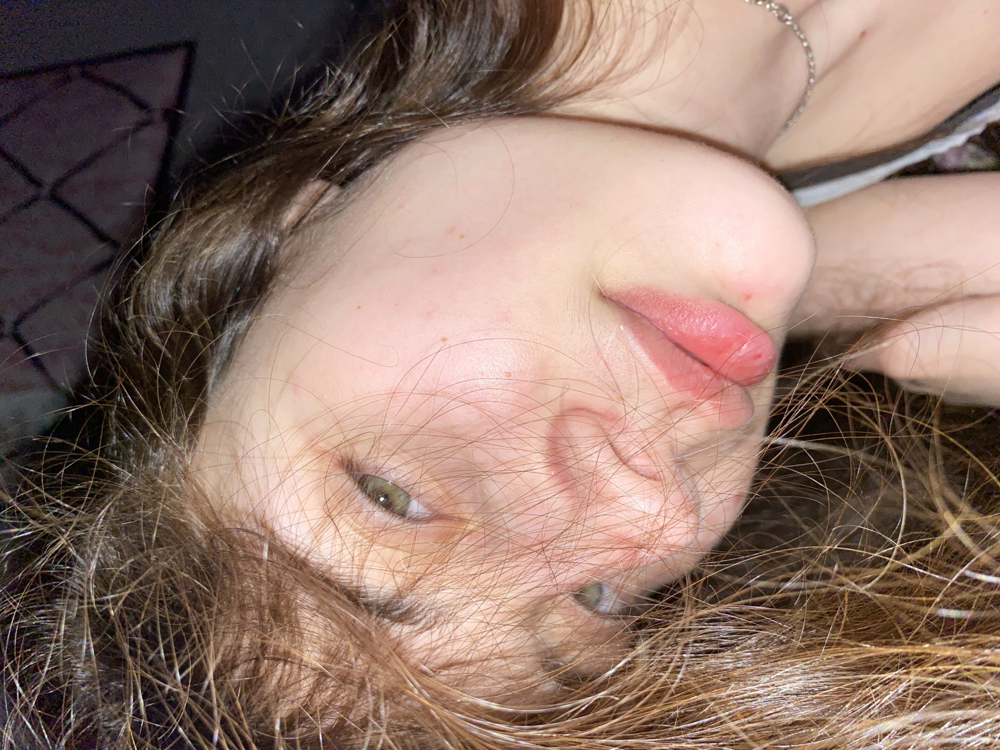 This close-up portrait, presented from an inverted perspective, features a young person with light brown or auburn hair and fair skin. The subject's green eyes are open and directed towards the camera, and their lips are naturally full, pink, and slightly parted. Scattered strands of hair frame their face and extend across the foreground.
Visible details include minor red blemishes on the person's forehead, cheek, and near the nose/mouth area. A thin silver chain necklace is worn around the neck/collarbone. The background is dark and dimly lit, with a faint, dark geometric pattern, possibly from a bed frame or headboard, visible in the upper left quadrant. The lighting suggests an indoor setting, potentially at night or in low light conditions. The overall impression is casual and relaxed, captured perhaps as a selfie while lying down. No text is visible in the image. The location context provided for this scene is Votkinsk, Russia.