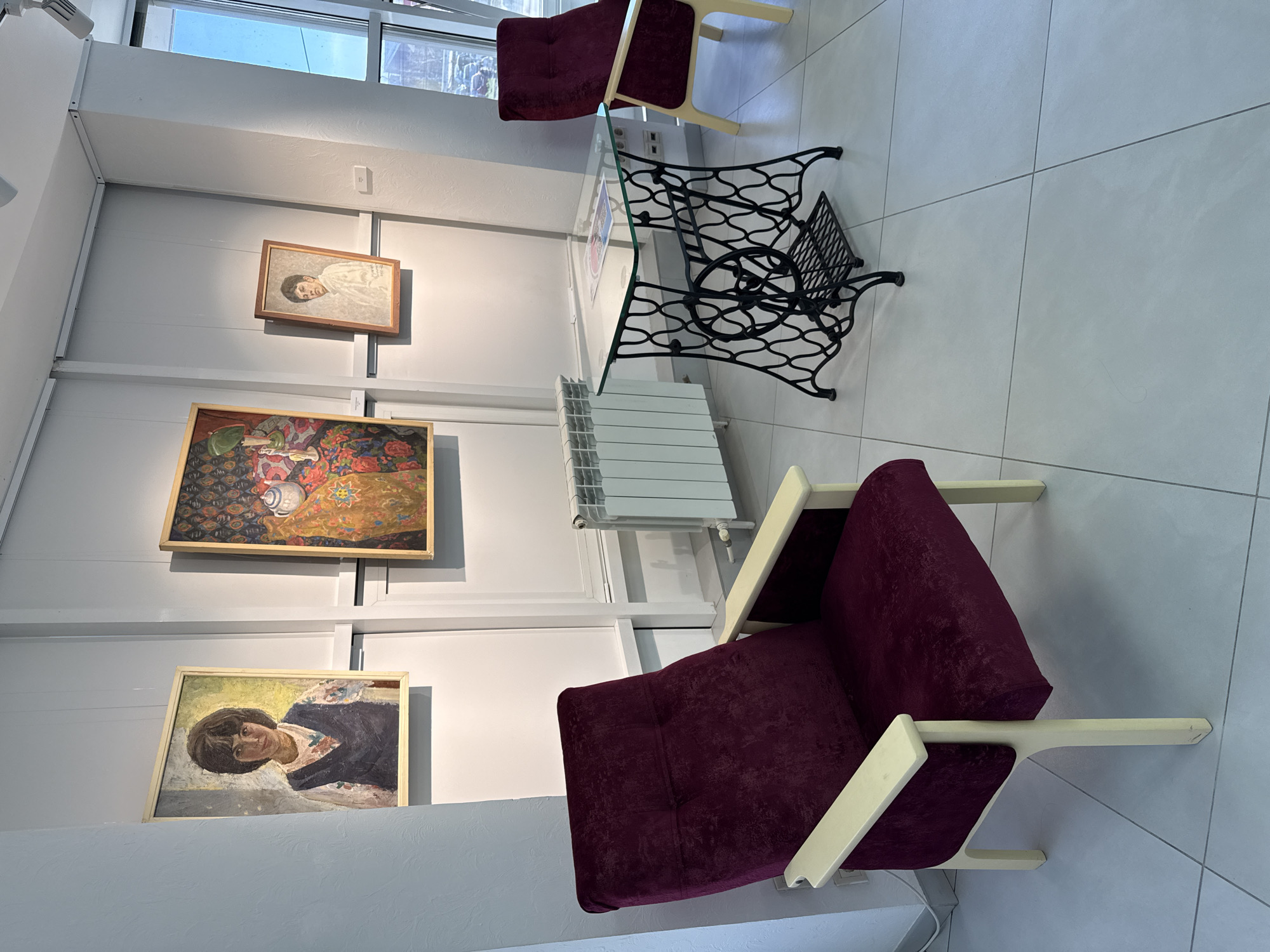 The image captures an interior scene, possibly an art gallery or exhibition space. Three framed paintings hang on a white wall, vertically aligned. The top painting appears to be a portrait of a person in light clothing. Below it, a painting displays a still life with a white vessel, accompanied by colorful geometric patterns and fruit. The bottom painting seems to feature another portrait.
In the foreground, two purple velvet armchairs with light-colored wooden frames are positioned. Between the armchairs and closer to the wall is a table with a glass top and a wrought-iron base. A white radiator is attached to the wall between the table and the lower armchair.
The floor is covered with large, light-colored tiles. The room is well-lit, and a window is visible in the top-left corner, suggesting natural light. The location is identified as Izhevsk, Russia. No people are present in the frame.