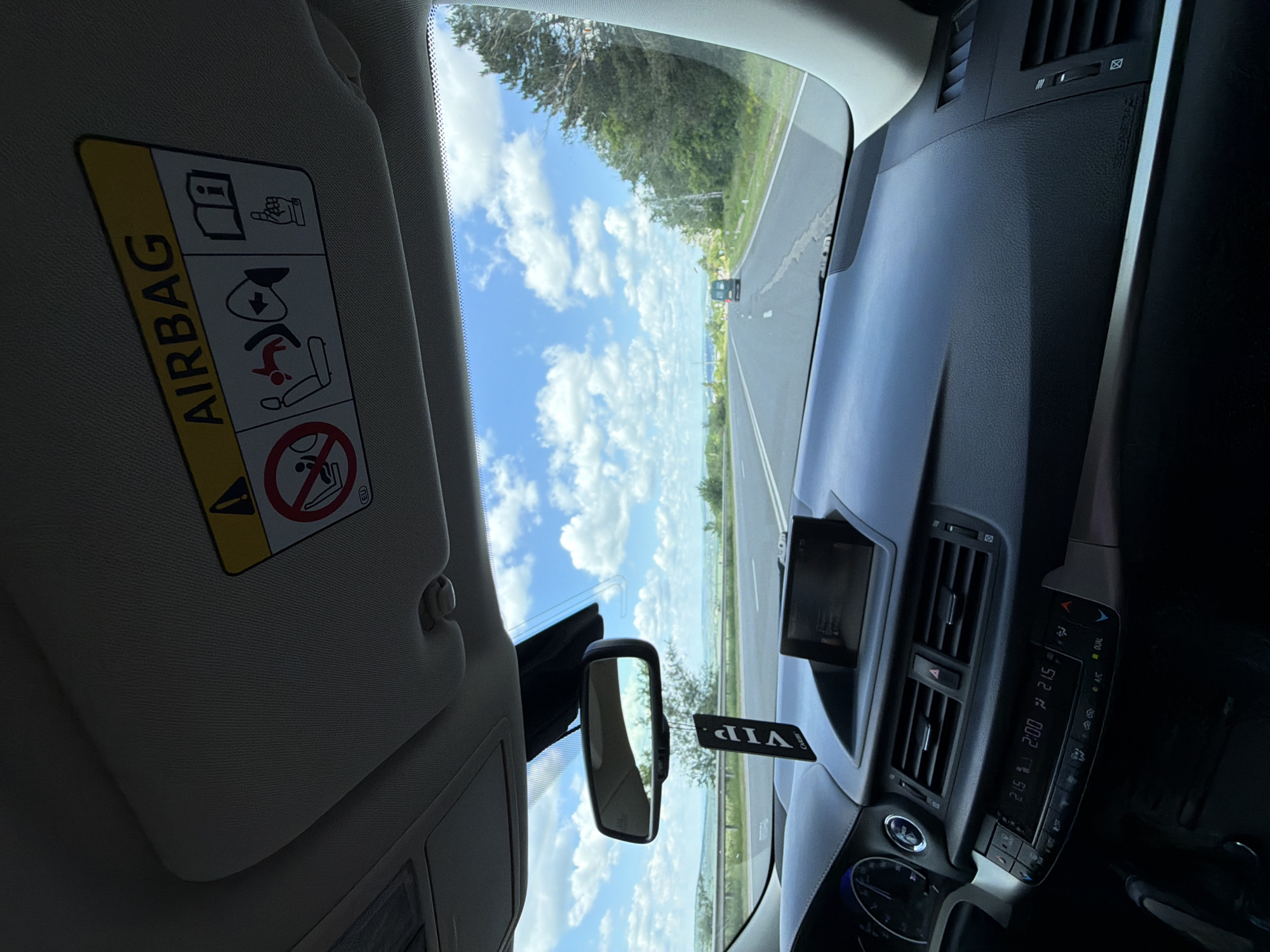 The image is taken from the interior of a car driving on a road. The perspective is from the driver's seat looking forward through the windshield. The dashboard is visible in the foreground, with a rear-view mirror hanging from the ceiling. A "VIP" air freshener hangs from the mirror. The sun visor is also visible, with an "AIRBAG" warning label. The road ahead is paved and appears to be a highway, with trees and vegetation on either side. The sky is blue with scattered white clouds. A dark-colored van is visible in the distance, driving in the same direction as the car. The image is likely taken during daylight hours. There are no people clearly visible in the image. The location of Peresecina, Moldova is not directly discernible from the image itself, as there are no identifying landmarks or signs.