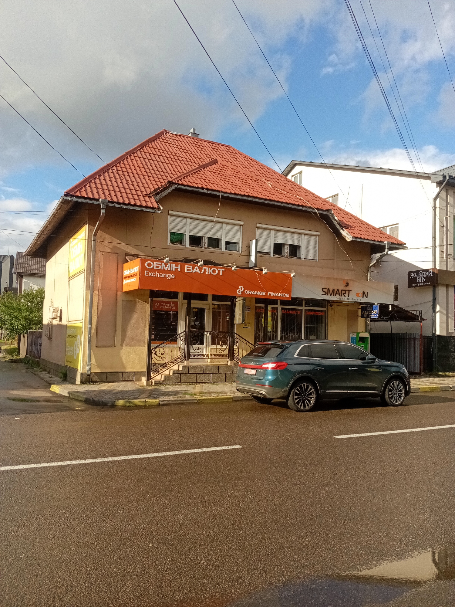 The image shows a street scene in Tiachiv, Ukraine. In the foreground, a dark teal SUV is parked on the right side of the road. The road is wet and has a white lane marking. In the background, there is a two-story building with a red tile roof. The building has several storefronts, including "ОБМІН ВАЛЮТ Exchange" (Currency Exchange) and "SMART N" which sells electronics. A bright orange sign with "ORANGE FINANCE" is visible above one of the storefronts. The sky is partly cloudy with patches of blue. The overall impression is of a commercial street in a European town.