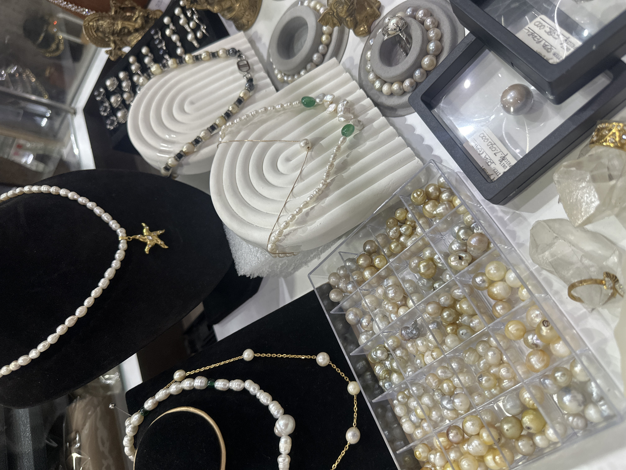 The image captures a detailed display of various pearl jewelry and loose pearls, likely in a retail setting such as a shop or market stall in Kuta, Indonesia. No people are visible.
The scene is dominated by a diverse collection of pearls. Several pearl necklaces and bracelets are arranged on black velvet display busts, white ribbed wave-like stands, and grey circular stands. One prominent necklace on a black bust features white baroque pearls with a gold-colored starfish pendant. Other necklaces incorporate emerald green beads or dark accent beads, alongside gold chains.
A large, clear plastic organizer box, divided into numerous compartments, is filled with an assortment of loose pearls. These pearls vary widely in size, shape (including round and baroque), and color, showcasing shades of cream, gold, white, and silver. Two black square display boxes are also present; one highlights a single dark, spherical pearl, while the other shows a white label. To the right, a gold ring adorned with clear gemstones rests upon a piece of clear quartz. The background features other out-of-focus jewelry items and possibly small decorative figurines.
The activity depicted is the showcasing of pearl jewelry and loose pearls, presumably for sale, customization, or exhibition to customers. The display is well-organized with bright, even lighting, emphasizing the luster and variety of the pearls. Visible text on the labels inside the black square display boxes includes "ITEM: PEARL" and "ITEM: 7.000.000," the latter likely indicating a price of 7 million, possibly in Indonesian Rupiah given the location context of Kuta, Indonesia.
