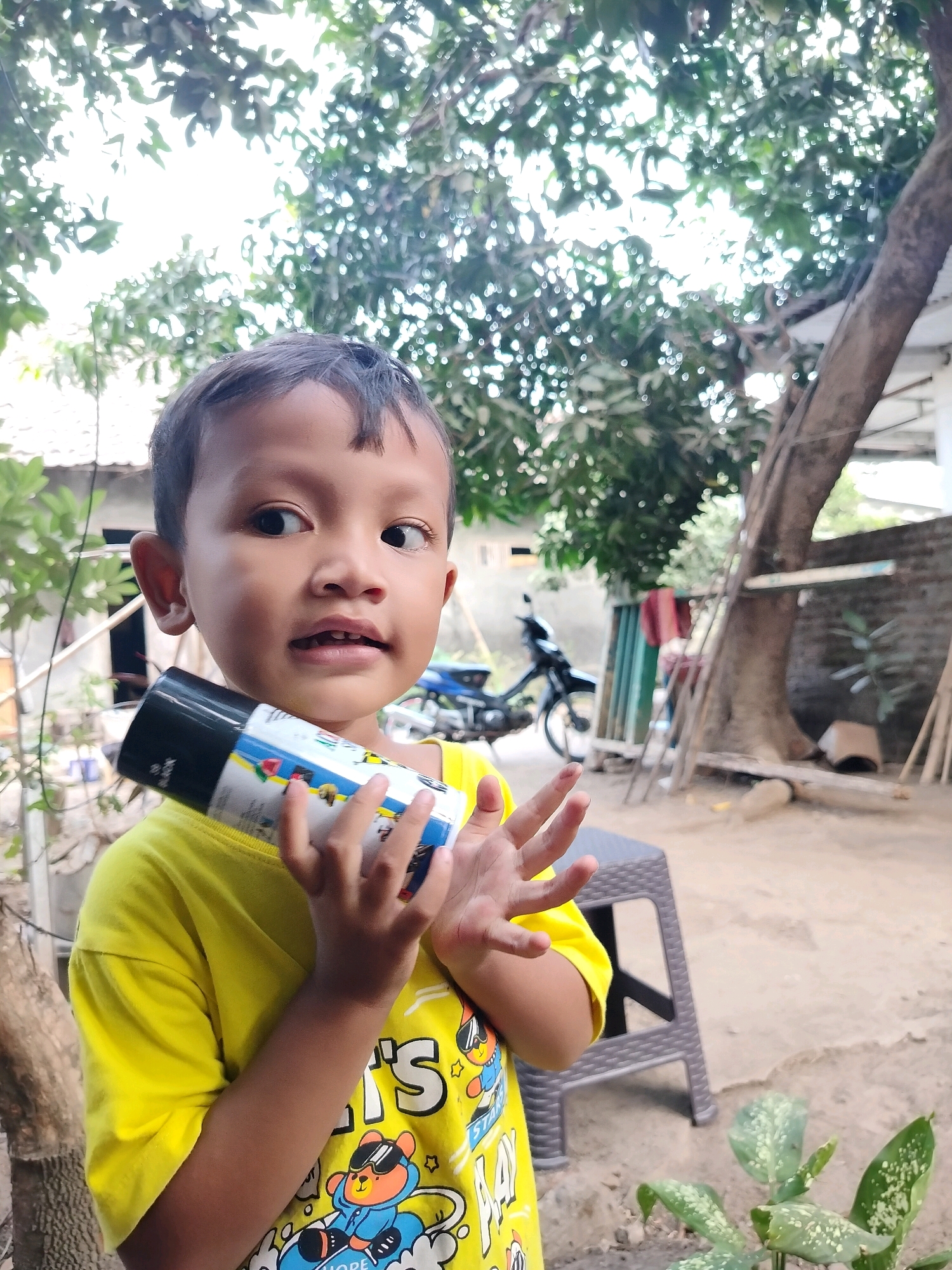 A young child, appearing to be around 4-5 years old, stands outdoors, holding a black and white spray can. The child has dark hair and is wearing a bright yellow t-shirt with cartoon characters on it. Their left hand is holding the spray can, while their right hand is open, fingers extended. The background suggests a rural or suburban setting, with trees, a motorcycle parked behind the child, and what appears to be a brick wall and some sort of structure in the distance. The lighting is bright, indicating it's daytime. The setting is identified as the city of Beloh, Indonesia.