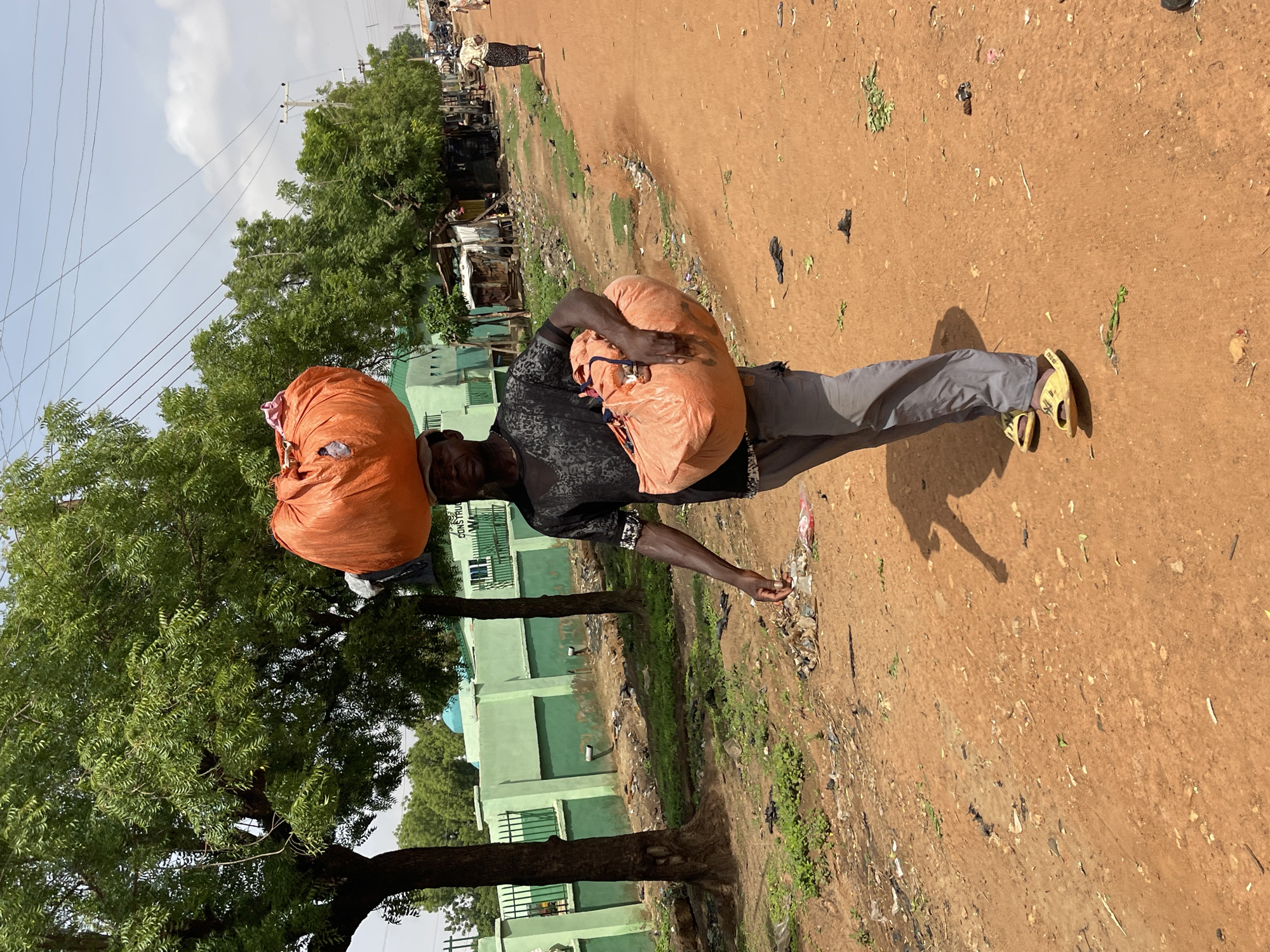 In Muchiya, Nigeria, an adult Black male balances two large orange bags on his head while walking along a dirt road. He is wearing grey pants, a dark printed shirt, and yellow sandals. The road is strewn with litter. Behind him are trees and a green building with a blue dome visible in the distance. A person walks in the background. Power lines are overhead.