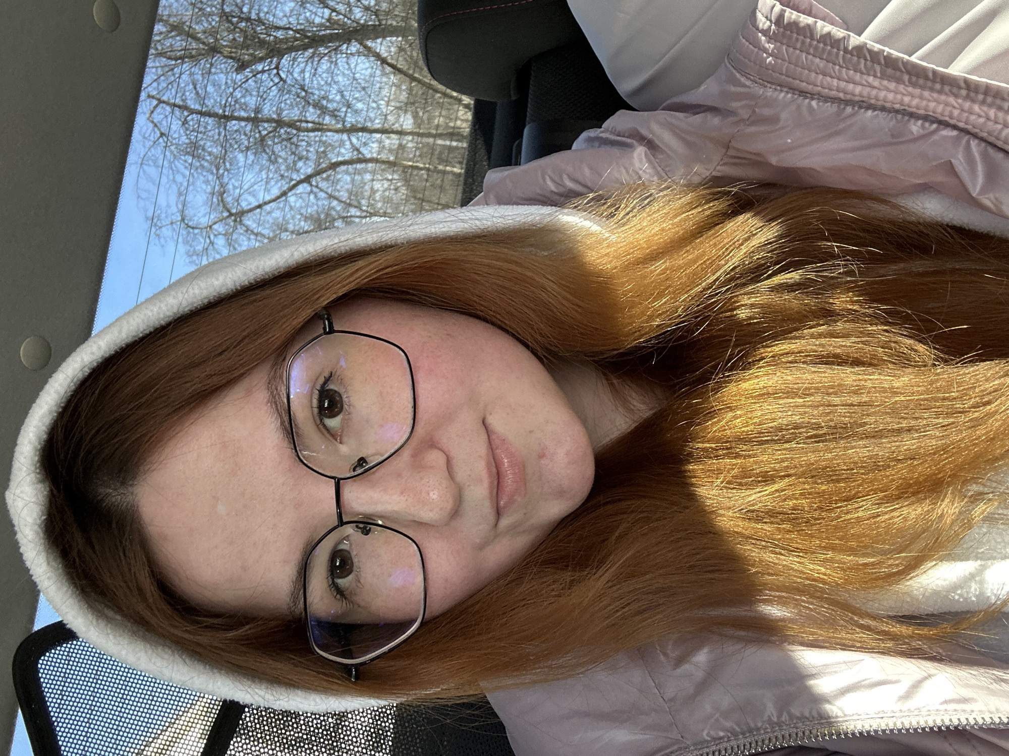 A person with red hair and glasses, wearing a white hooded sweatshirt and a light pink jacket, is in a vehicle. The view through the vehicle window shows trees and a bright sky. The specific location is undeterminable from the image.