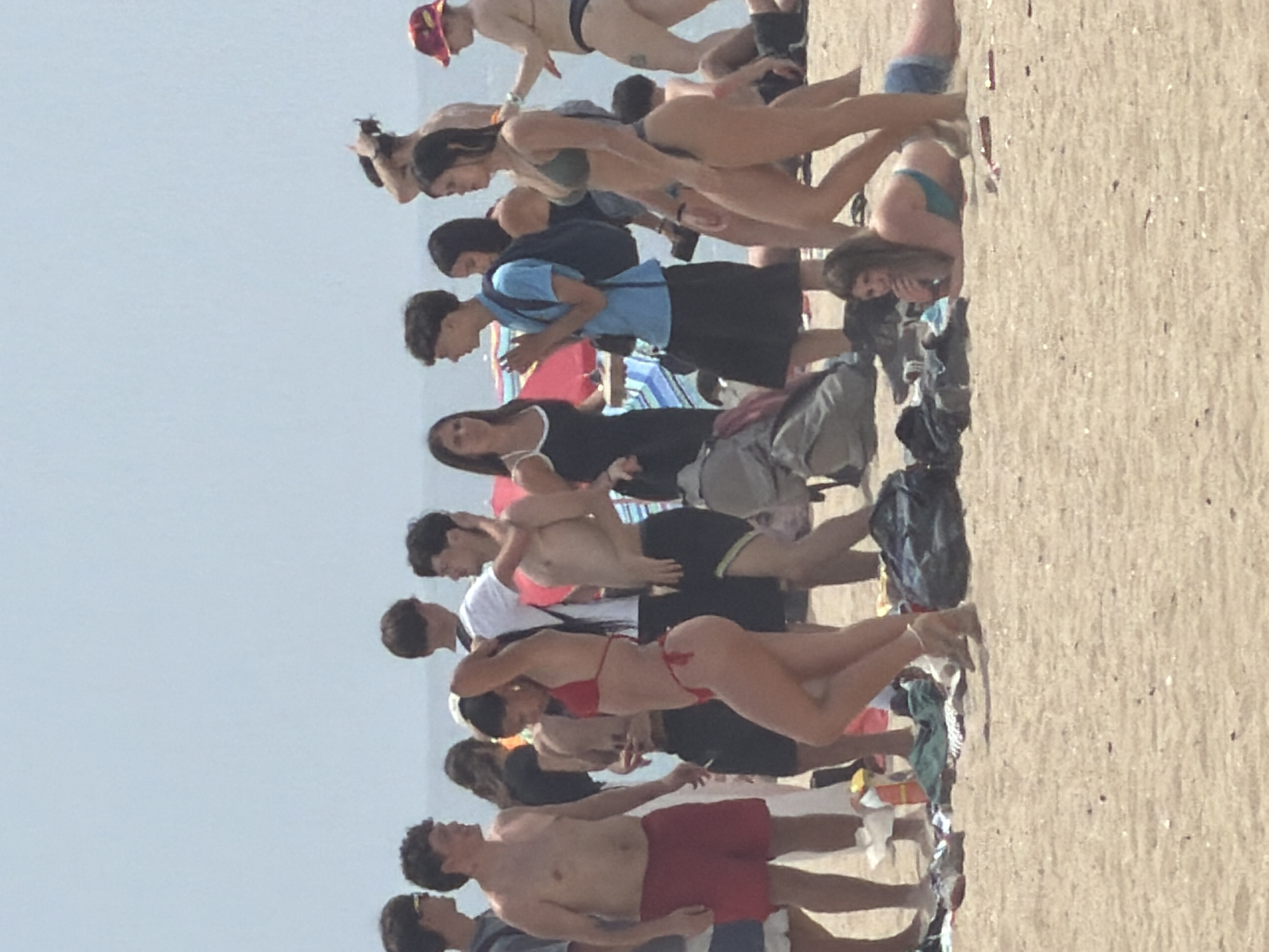 A group of people are gathered on a beach in Espinho, Portugal. The sky is a clear, pale blue, and the sand is a light tan color. In the foreground, a woman in a red bikini is posing on the sand, with her right hand on her head and her left hand on her hip. To her left, a man in red shorts stands with his arms crossed. Behind them, several other individuals are seen in various states of dress and activity. Some are talking, while others are looking out at the ocean. There are beach towels and bags scattered on the sand. In the background, more people and beach umbrellas are visible. The overall scene depicts a casual day at the beach.