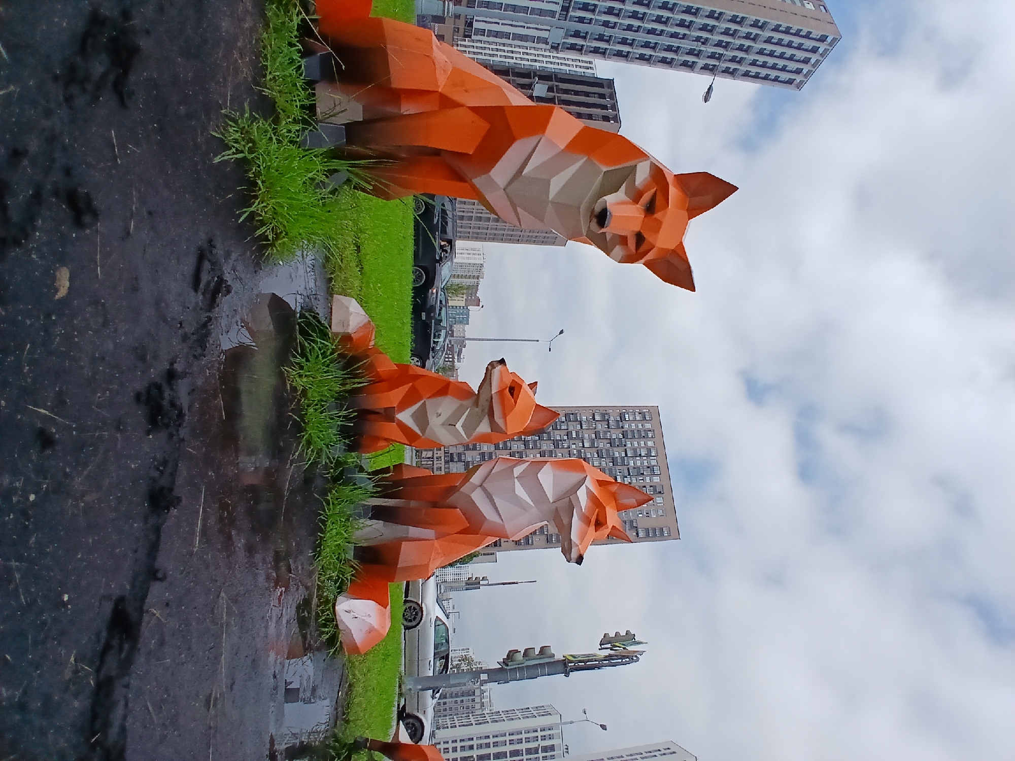 Three abstract, polygonal fox sculptures, rendered in orange and white, are positioned on a patch of green grass. The largest fox is on the left, looking directly at the viewer. A medium-sized fox sits to its right, facing left. A smaller fox is in front of the medium-sized one, also facing left. Behind the sculptures, a grey asphalt surface with puddles of water reflects the overcast sky. Several modern high-rise buildings, characteristic of Yekaterinburg, Russia, dominate the background. Traffic lights and streetlights are visible. Two dark-colored cars and a silver sedan are parked or driving on the road in the middle ground. The sky is filled with white and grey clouds.
