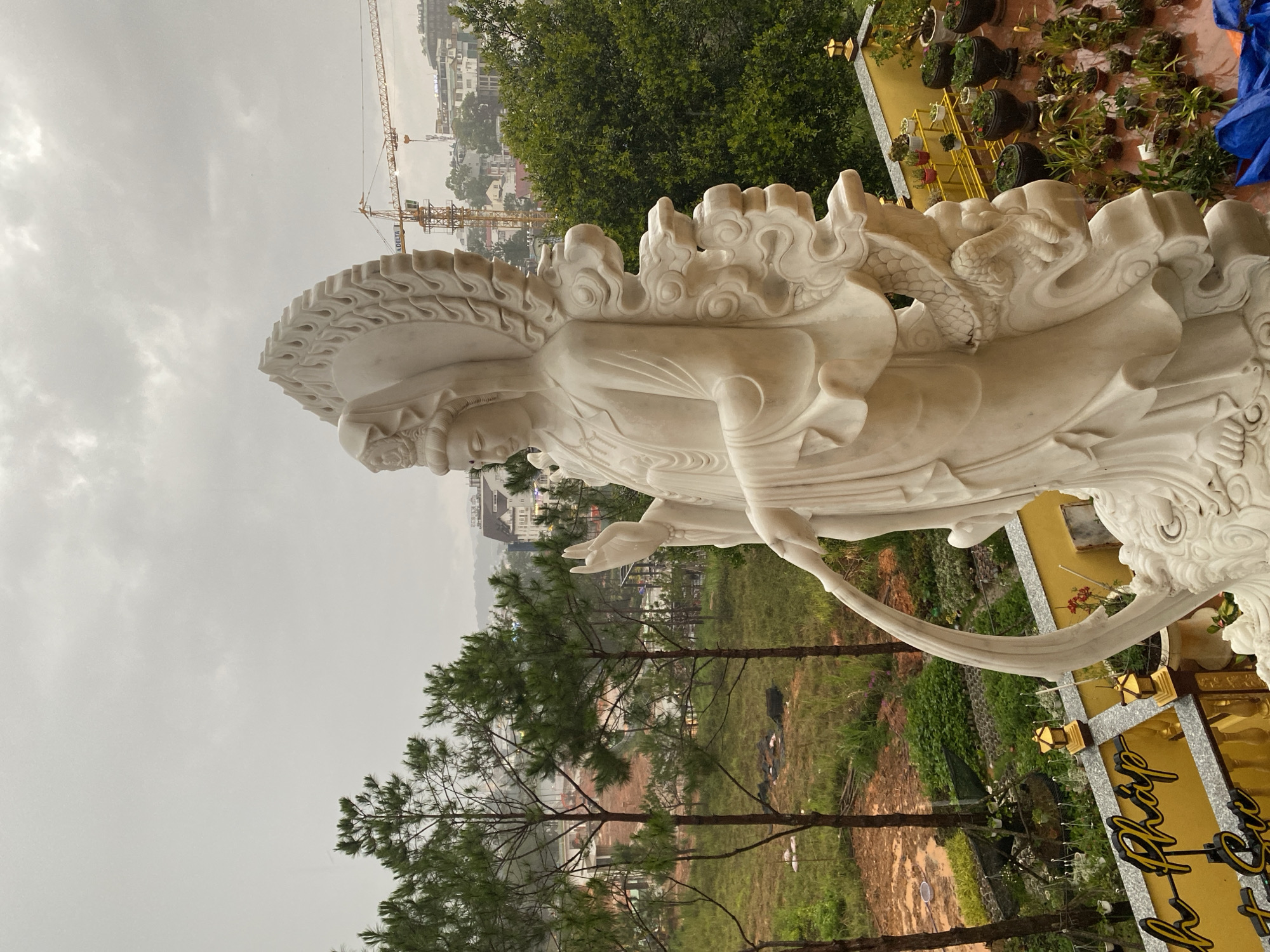 The image depicts a white marble statue, possibly of a deity or revered figure, located in Da Lat, Vietnam. The statue is intricately carved, with detailed features on the figure and ornate decorations around it, including what appears to be a dragon figure on the right side of the image. The statue is set against a backdrop of green trees, vegetation, and a sloped landscape. Buildings and construction cranes are visible in the distance, suggesting the statue is located on the outskirts or high point within the city. The foreground shows pine trees. The sky is overcast, indicating possible rain. There's a gold-colored sign with black lettering that is partially obscured. The statue appears to be the primary subject of the image.