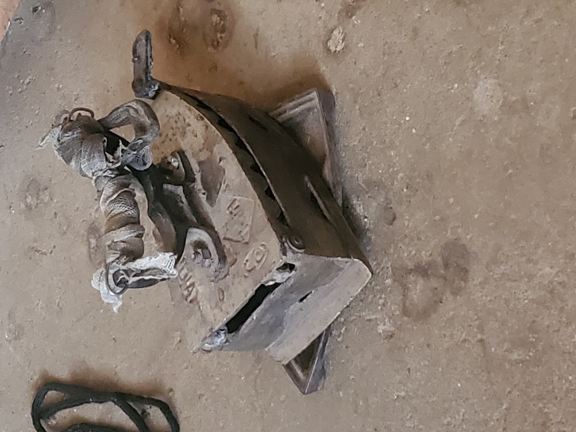 An antique cast iron flat iron sits on a metal stand on a dusty, concrete-like surface. The iron is a dull metallic gray, showing signs of age and wear, with some rust and discoloration visible. A piece of dirty, white cloth is wrapped around the handle and a metal loop attached to the top of the iron. A thick black cord is coiled to the left of the iron. The background is a rough, textured surface that appears to be dirt or concrete, with small pebbles and debris scattered across it. The lighting is somewhat dim, casting soft shadows. The image was taken in Kabwir, Nigeria.