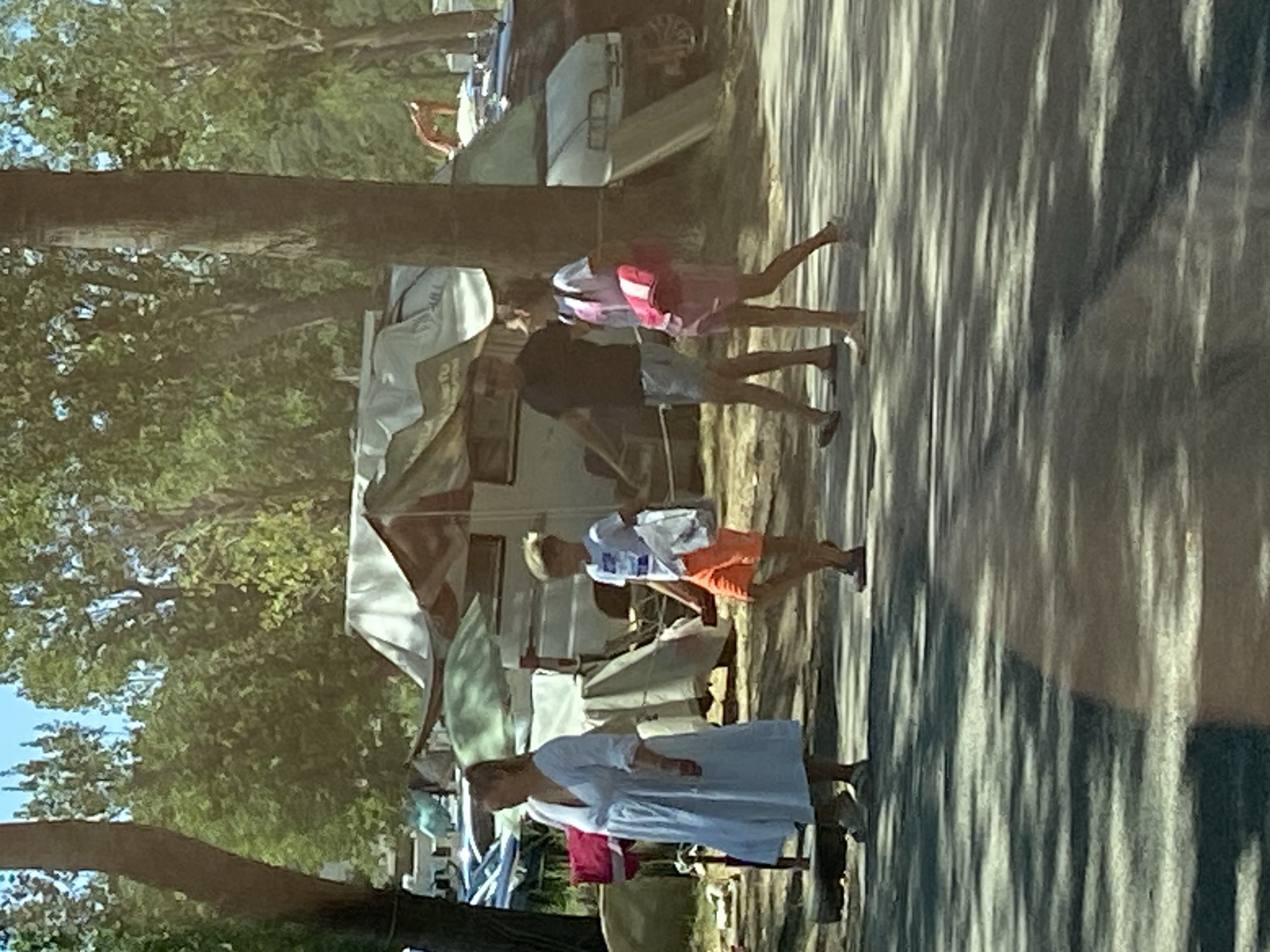 In Prijevor, Montenegro, on a sunny day, a family walks along a paved path through a wooded area with a campsite. A woman in a long white dress walks left to right, holding a pink bag in her left arm. To her right, a boy in an orange t-shirt and shorts walks left to right, carrying a white paper bag in his right hand. Behind them, a man in a dark t-shirt and shorts walks left to right. To his right, a young girl in a pink t-shirt and skirt walks left to right, carrying a pink bag over her left shoulder. In the background, several white tents and campers are visible among the trees.