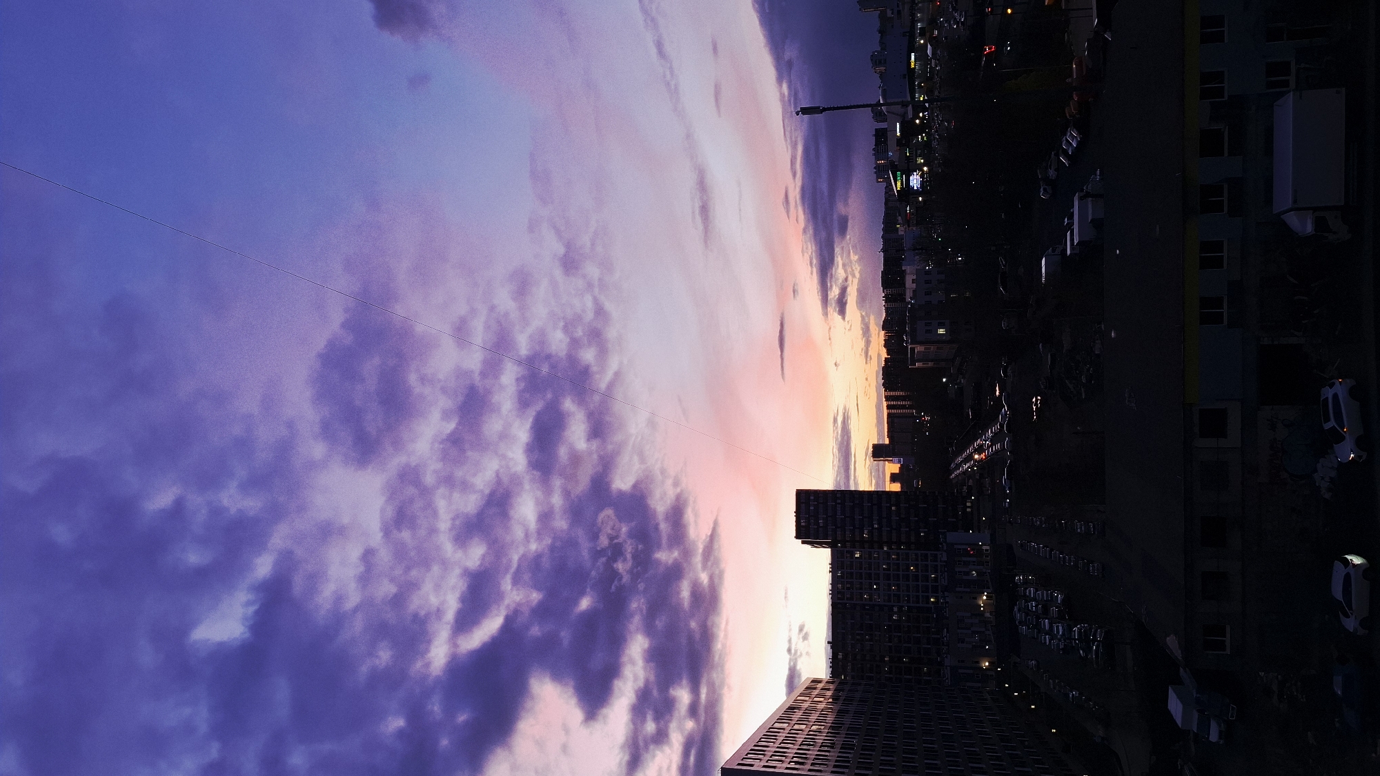 The image captures a vibrant cityscape at sunset or twilight, viewed from an elevated perspective. The scene is dominated by a dramatic sky, painted with gradients of deep purple and blue on the left, transitioning through various shades of lavender and pink, to bright orange and yellow hues near the horizon on the right. Numerous clouds, varying in shape and density, are scattered across the sky, reflecting these brilliant colors.
Below the colorful sky, the urban landscape stretches horizontally, mostly silhouetted against the declining light. A collection of buildings, primarily residential or commercial high-rises, are visible. Many windows in these buildings are illuminated, casting warm glows against their dark facades. On the ground level, streets are lined with parked vehicles, and faint lights suggest ongoing activity. Some streetlights and distant city lights contribute to the artificial illumination of the scene.
Thin, dark wires or power lines stretch diagonally across the upper and middle sections of the sky, adding a man-made element to the natural spectacle. The overall impression is one of a serene yet active urban evening. The time of day is clearly sunset or late twilight, evidenced by the rich sky colors and the mix of natural and artificial lighting. No specific geographical landmarks or discernible text are visible within the image itself to identify the location as Saint Petersburg, Russia.