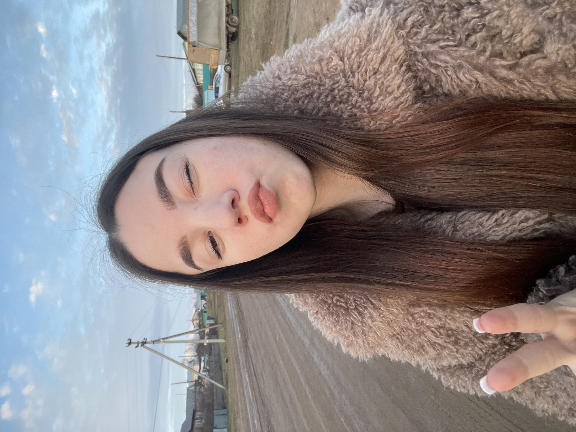 A young woman with long, straight dark brown hair is featured in a vertical selfie, taken outdoors. Her face is in the foreground, with her eyes partially closed and her lips pursed in a stylized "kissy face" expression. She has well-groomed eyebrows. She is wearing a fluffy, textured coat in a light brown or beige color, visible across her chest and shoulder. Her right hand, showing a French manicure with white-tipped nails, is visible in the lower right quadrant of the image.
The background shows an expansive outdoor scene under a partly cloudy sky. The sky is a soft blue with diffuse white clouds, suggesting natural daylight, possibly during the late afternoon or early evening. The ground beneath the sky appears to be an unpaved or dirt road extending into the distance. Several utility poles with wires line the side of the road, and in the background, a few modest buildings or structures are visible. A vehicle, possibly a truck or car, can be faintly seen in the far distance near some buildings. The overall setting suggests a rural or semi-rural area, consistent with the given location context of Ольховка, Russia. There is no visible text in the media file.