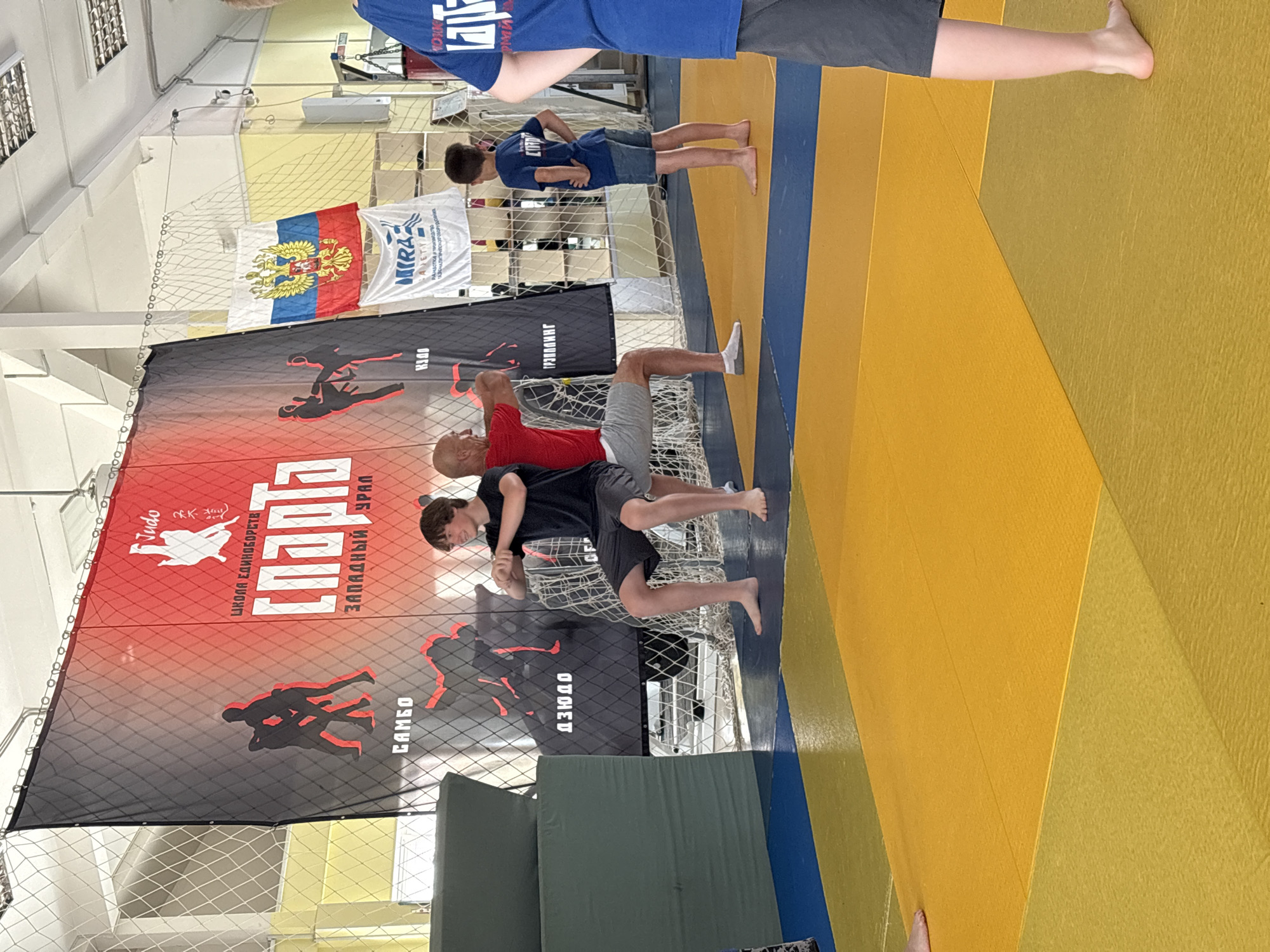 In a gymnasium in Chaikovsky, Russia, a youth wrestling event is underway. A large banner with graphics of judo fighters hangs behind a young boy in a black t-shirt and shorts performing a squat. To his right, an adult male in a red t-shirt and grey shorts demonstrates a similar stance, with his hands extended. A younger boy in a blue t-shirt and denim shorts observes the demonstration. The gymnasium floor is covered with yellow and blue mats. Russian flags and a banner with the Mirazh company logo are visible in the background.